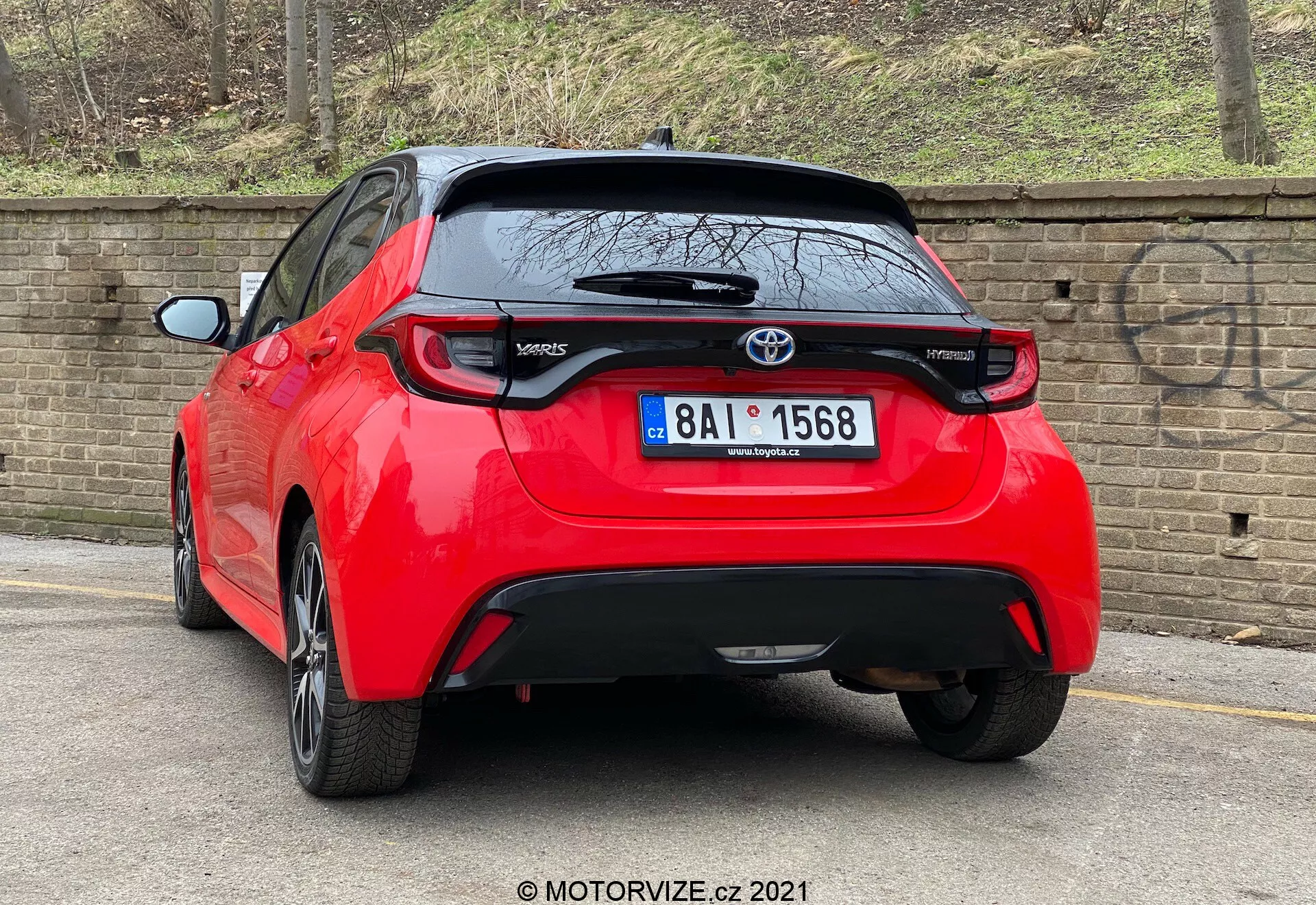 Un'immagine ad alta risoluzione di una Toyota Yaris 2020 rossa hatchback ripresa da un'angolazione posteriore di tre quarti. Il veicolo presenta luci posteriori distintive, un emblema Toyota posto centralmente sopra una probabile telecamera posteriore e il badge del modello 'Yaris' nella parte inferiore sinistra del portellone. Sotto l'area della targa c'è un pezzo di rifinitura nera che potrebbe essere di plastica o fibra di carbonio, con un piccolo spoiler posteriore che si estende sopra il lunotto. L'auto, parcheggiata vicino a un marciapiede su una strada pavimentata accanto a un muro di mattoni, suggerisce un'ambientazione urbana o residenziale.