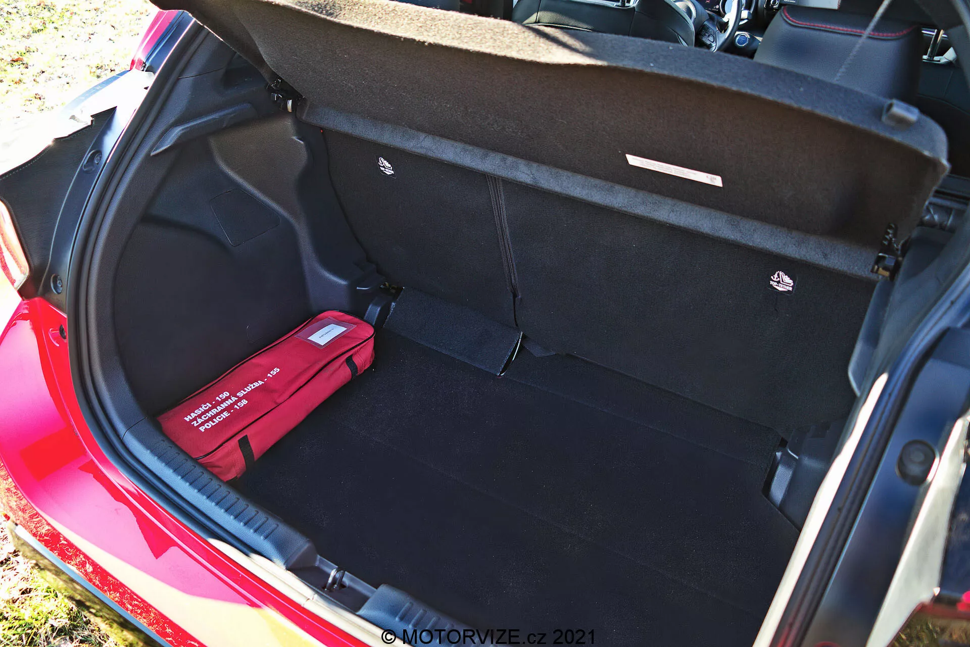 Bagagliaio di una Toyota Yaris 2020 rossa vista da un angolo elevato, che mostra l'interno pulito e spazioso con moquette nera, coperchio del bagagliaio aperto con maniglie di tiro, un pavimento di carico piatto, un gancio di carico a destra e un kit di pronto soccorso o di emergenza rosso a sinistra. Questa fotografia diurna evidenzia la grande capacità di stoccaggio e le caratteristiche organizzative del veicolo.