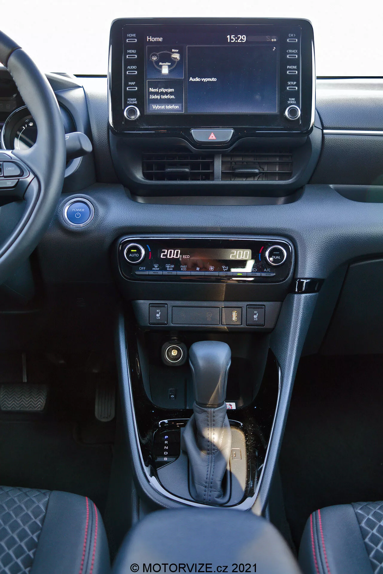 Vista interna di una Toyota Yaris 2020 con attenzione alla console centrale e agli elementi del cruscotto. La foto, scattata dal sedile passeggero anteriore, mostra un sistema di infotainment digitale touchscreen che visualizza una schermata principale con l'ora 15:29 e varie opzioni di menu, bocchette dell'aria centrali e comandi user-friendly per l'aria condizionata e il riscaldamento con un display digitale della temperatura impostata a 20,0 gradi. La console centrale presenta una leva del cambio rivestita in pelle con un accento argentato e un freno a mano manuale. Gli interni sono progettati con toni scuri e finiture metalliche, offrendo un'estetica moderna e sofisticata.