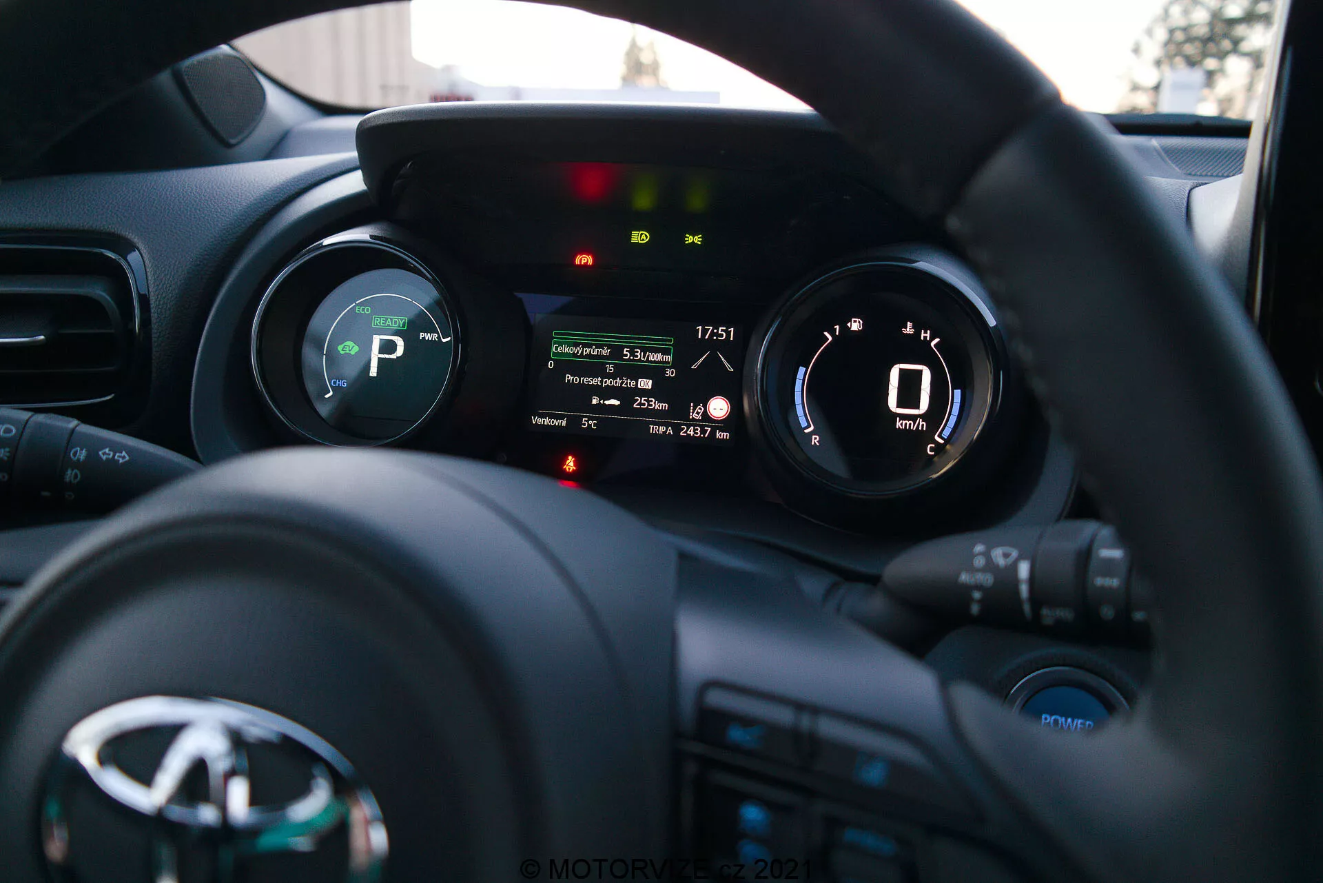 Una fotografia dell'interno della Toyota Yaris 2020 dalla prospettiva del conducente, con messa a fuoco sul volante e sul quadro strumenti. Il volante, con il logo del produttore e vari pulsanti di controllo per media, comunicazione e controllo della velocità, è in basso. Il quadro strumenti dietro il volante presenta un display digitale che mostra un tachimetro a 0 km/h e indicatori per la marcia (P per Park), promemoria delle cinture di sicurezza, allarme porta aperta e altre luci di stato del veicolo. La bassa profondità di campo mette in evidenza il quadro strumenti, mentre il cruscotto e l'interno dell'auto sono leggermente sfocati sullo sfondo. L'immagine, probabilmente scattata nel tardo pomeriggio o prima serata, mostra una strumentazione moderna e tecnologia di interfaccia del veicolo.