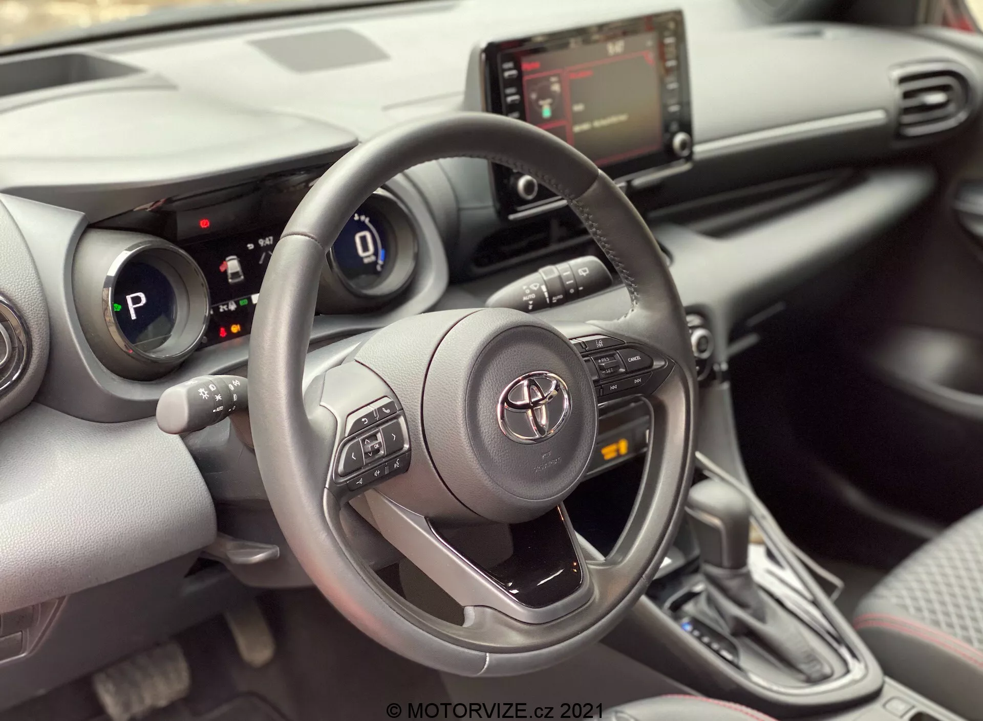 Vista degli interni di una Toyota Yaris 2020 dal sedile del passeggero anteriore, concentrandosi sull'area del conducente. L'immagine mette in evidenza un volante Toyota a tre razze con pulsanti di controllo per audio, Bluetooth e cruise control, con il logo Toyota al centro. Dietro il volante, il quadro strumenti digitale è parzialmente visibile indicando che l'auto è in modalità 'P' (park). La console centrale mostra lo schermo dell'infotainment e i controlli HVAC sotto di esso, insieme a possibili porte USB o ausiliarie. L'immagine mostra anche una leva del cambio automatico con un design contemporaneo e copertura simile al cuoio. Gli interni presentano un design moderno con una combinazione di materiali morbidi al tatto e accenti argentati, insieme a porta bicchieri o piccoli compartimenti di stoccaggio accanto alla leva del cambio. Un pannello della porta parzialmente visibile rivela un pannello di controllo delle finestre, suggerendo finestrini elettrici.