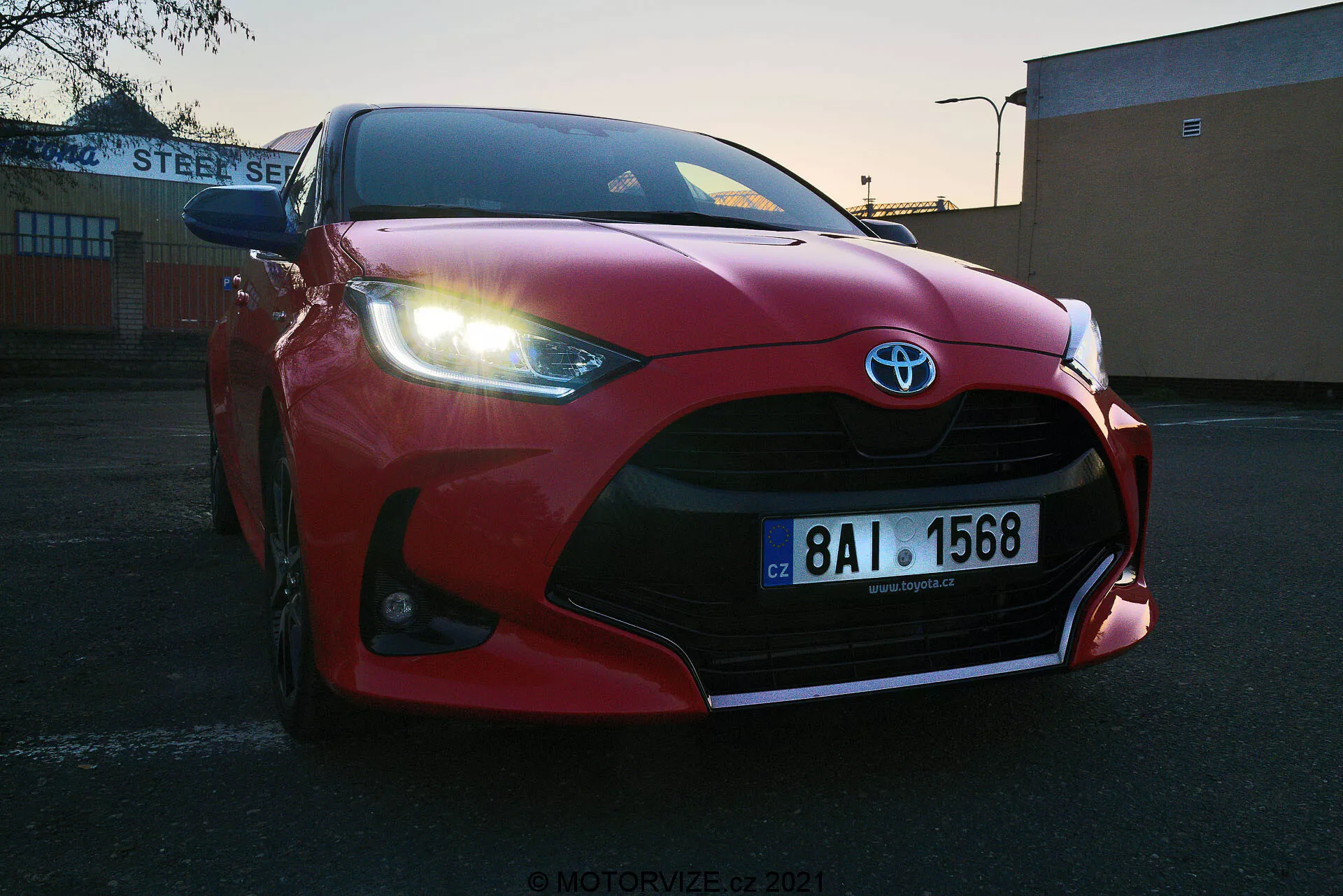 Primo piano a bassa angolazione di una Toyota Yaris 2020 rossa parcheggiata all'aperto all'alba o al tramonto. Griglia anteriore, paraurti, cofano scolpito aerodinamicamente, fari LED affilati e grandi prese d'aria che evidenziano il distintivo Toyota. Targa europea visibile, situata contro lo sfondo industriale o commerciale dell'edificio 'STEEL SERVICE'. Cielo limpido con luce sfumata che mette in risalto il design sportivo del veicolo.