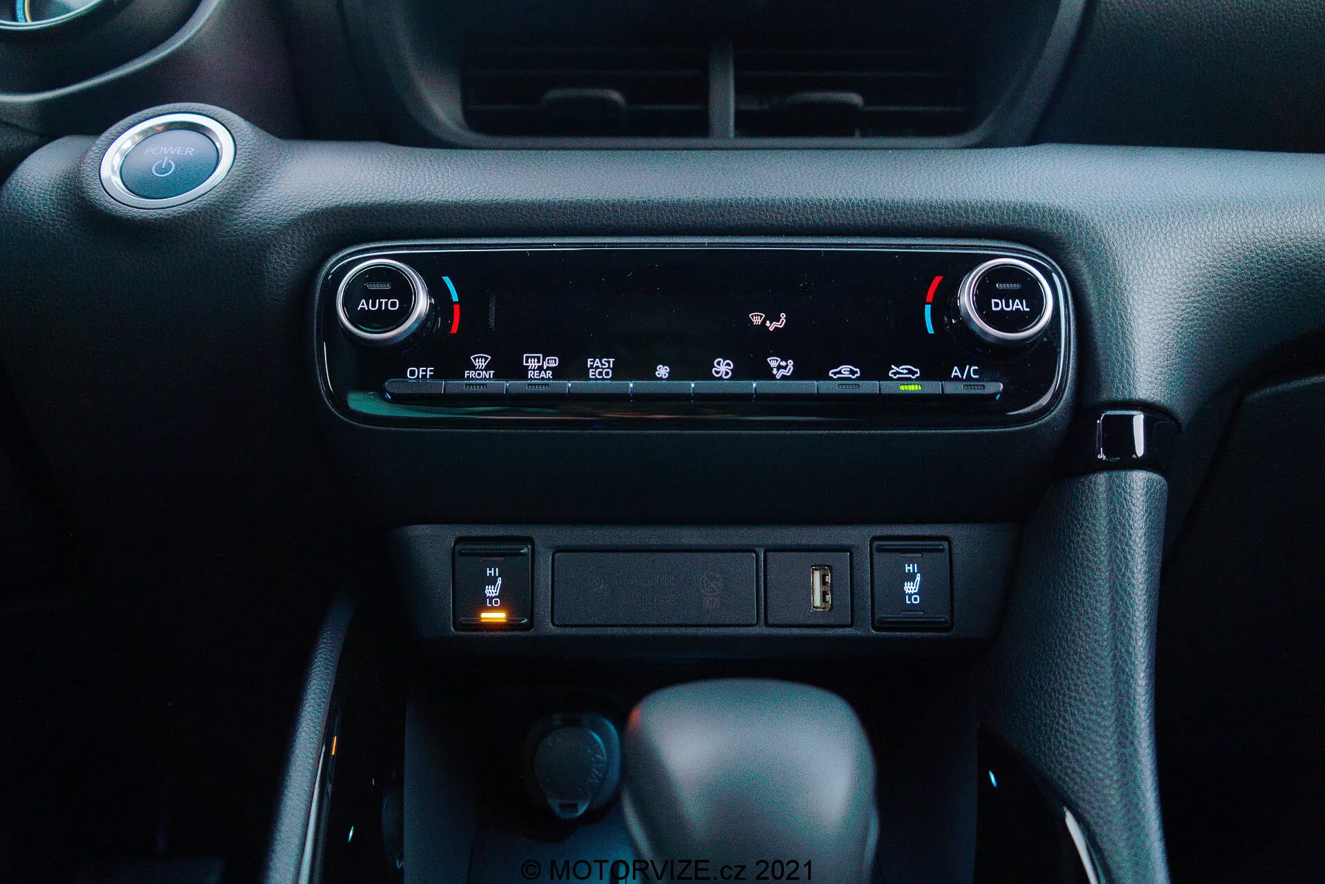 Vista ravvicinata del cruscotto della Toyota Yaris 2020: L'immagine evidenzia il sistema di controllo del clima con climatizzazione automatica a doppia zona, con pulsanti 'AUTO' e 'DUAL' per regolazioni indipendenti della temperatura per il conducente e il passeggero. Sotto i controlli del clima, è visibile uno slot di archiviazione con una porta USB e una potenziale presa di corrente da 12V. Sotto questo, si vedono tre manopole: quella centrale è la leva del cambio in 'Park', indicando una trasmissione automatica, una manopola rotante più piccola a sinistra probabilmente per la selezione della modalità di guida, e un pulsante a destra che potrebbe essere un freno di stazionamento elettronico. Il materiale del cruscotto ha uno schema di colori scuri con una finitura strutturata premium, creando un ambiente moderno e intuitivo.