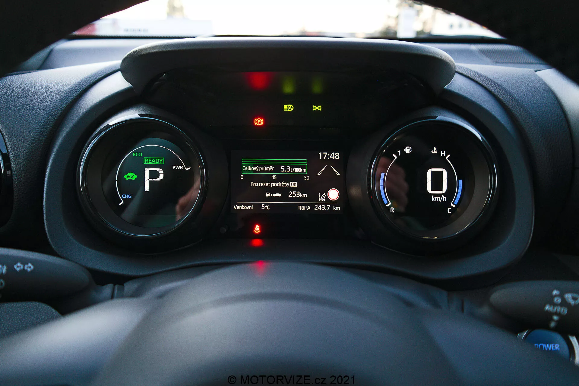 Vista interna del cruscotto della Toyota Yaris 2020 con un focus sul quadro strumenti, presa dalla prospettiva del conducente che mostra strumenti digitali, indicatori ibridi o elettrici e un display multifunzione con caratteristiche come distanza del viaggio, temperatura esterna e autonomia della batteria.