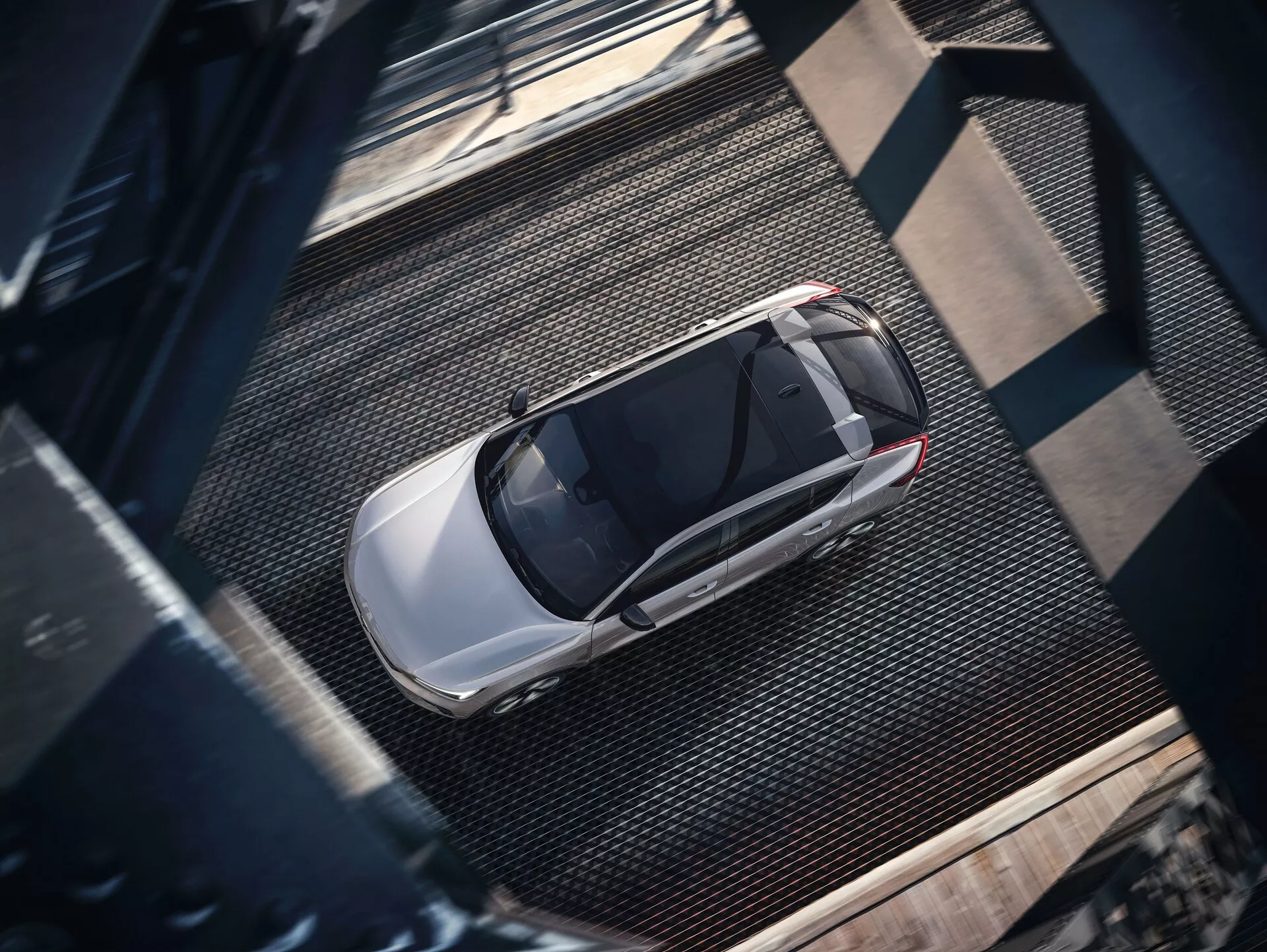 Vista aerea di una Volvo C40 2021 di prima generazione, catturata da una prospettiva dall'alto. L'immagine rivela la vista completa dell'elegante berlina moderna, caratterizzata da una finitura metallica lucida di colore chiaro, probabilmente argento o grigio chiaro. Le caratteristiche visibili includono il tetto, il cofano, il bagagliaio, i finestrini e parti delle porte, con un prominente tetto apribile che suggerisce una ventilazione avanzata. L'auto è parcheggiata su una superficie testurizzata in stile industriale con trame metalliche a griglia e travi rivettate scure, proiettando ombre che indicano una luce laterale probabilmente al mattino o nel tardo pomeriggio.