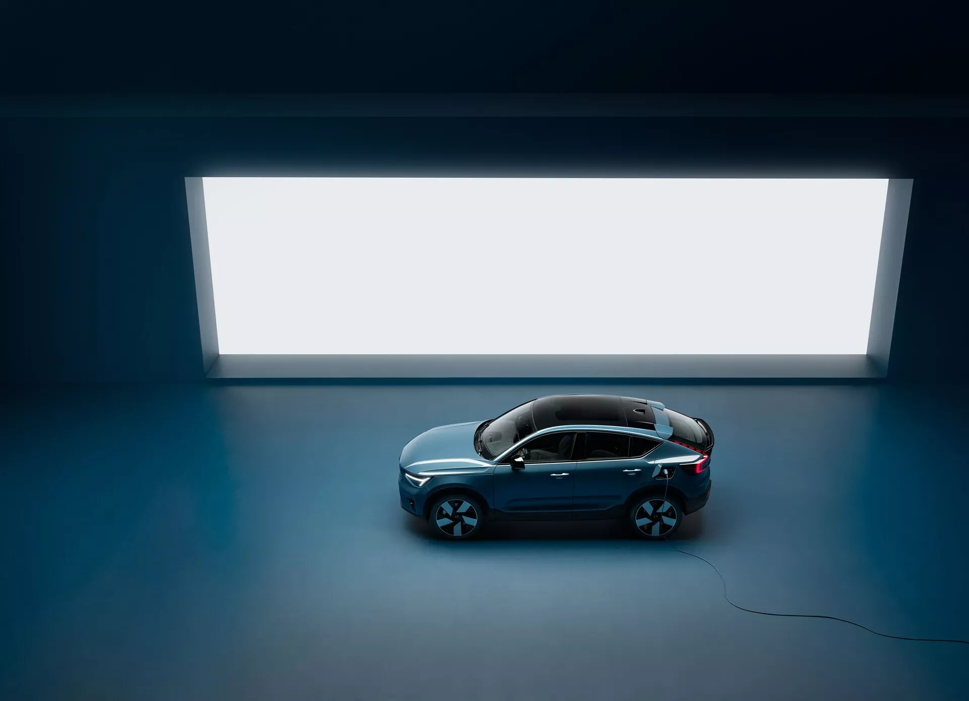 Inquadratura dall'alto di una Volvo C40 2021 berlina con quattro porte e tetto panoramico, vista dall'alto contro un pavimento scuro con un'ampia fonte di luce rettangolare luminosa sullo sfondo, che mostra un design pulito e minimalista con una carrozzeria di colore scuro e finitura lucida e grandi ruote in lega multiraggio, evidenziando la sua estetica lussuosa e moderna.