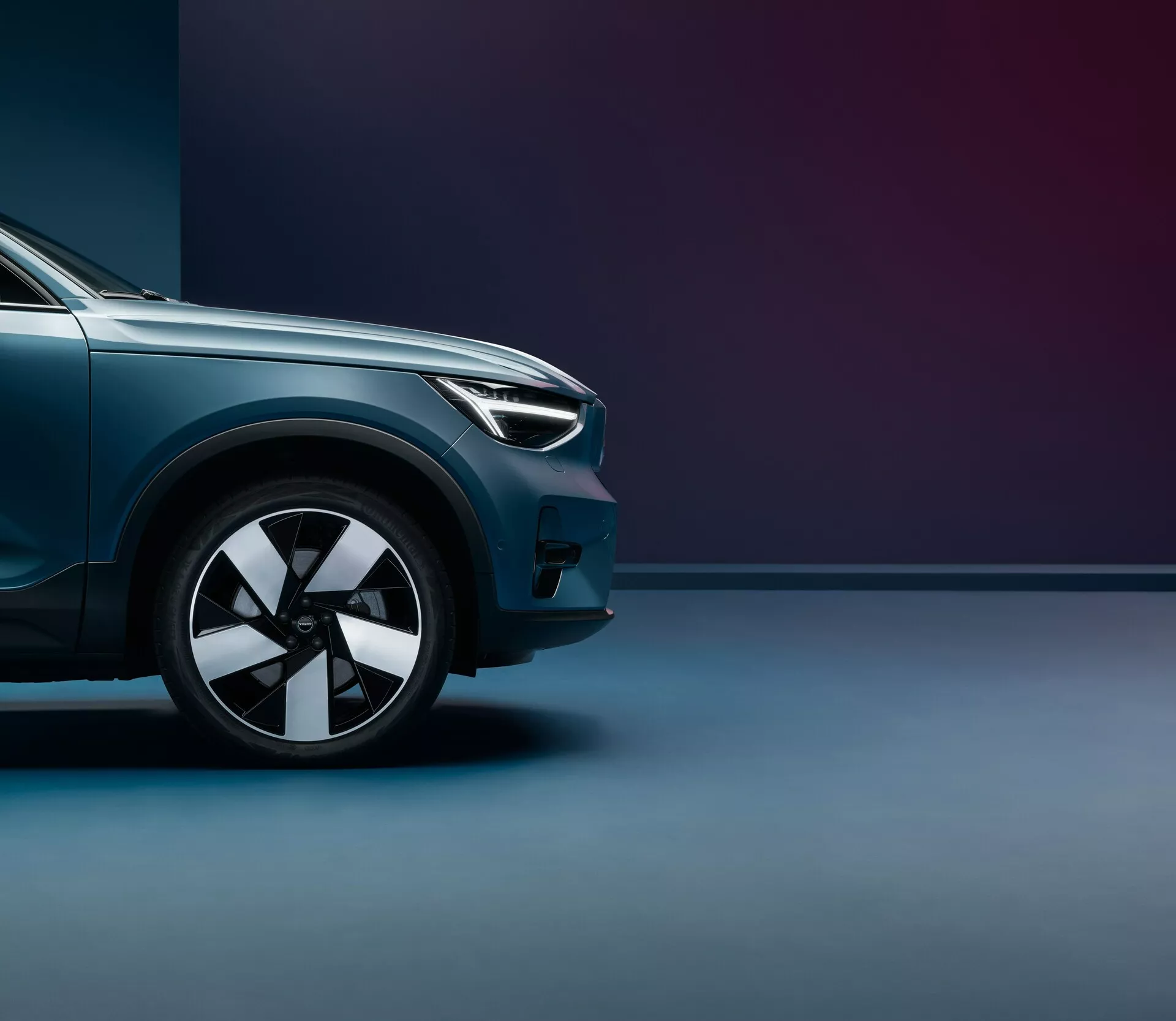 Scatto in basso angolato del Volvo C40 2021 (prima generazione), con attenzione al quarto anteriore dal pneumatico anteriore fino alla metà del cofano. Evidenziati il design moderno dei fari, il cerchio in lega a più raggi, la vernice metallizzata lucida e le linee aerodinamiche. Lo sfondo bicolore con illuminazione soffusa sottolinea la finitura premium e la posizione imponente del veicolo.