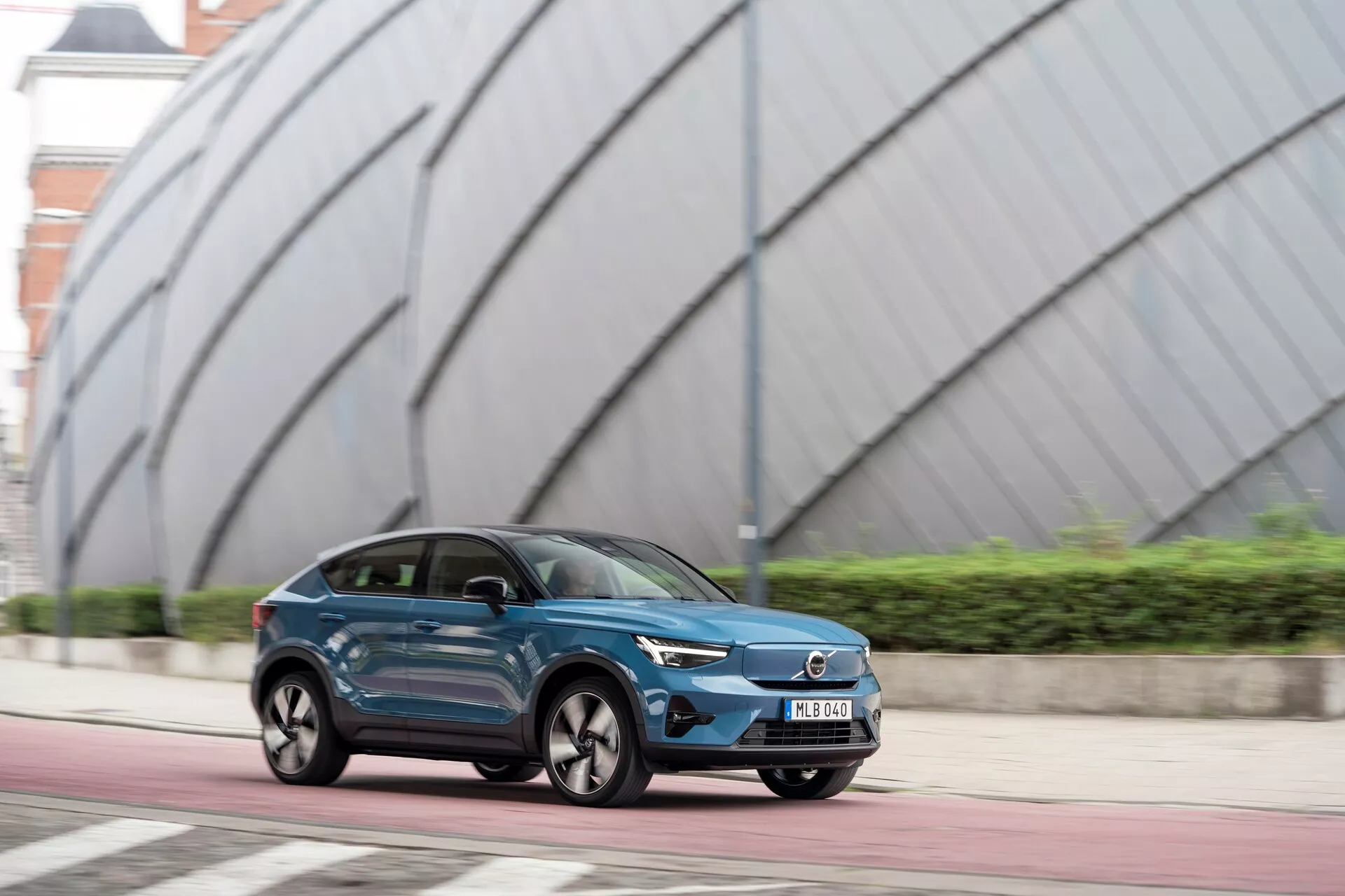Immagine di una Volvo C40 del 2021 (prima generazione) in movimento su una strada urbana. Il SUV crossover a quattro porte è catturato da un angolo di vista di tre quarti frontale, evidenziando linee eleganti e aerodinamiche, pannelli laterali sagomati e ruote distintive. La vernice metallica blu scuro e la griglia anteriore prominente con il logo del produttore suggeriscono un possibile modello elettrico o ibrido. I fari a LED con design stretto e aggressivo e le luci diurne visibili aggiungono al suo fascino moderno. Lo sfondo con architettura moderna enfatizza l'ambiente urbano.