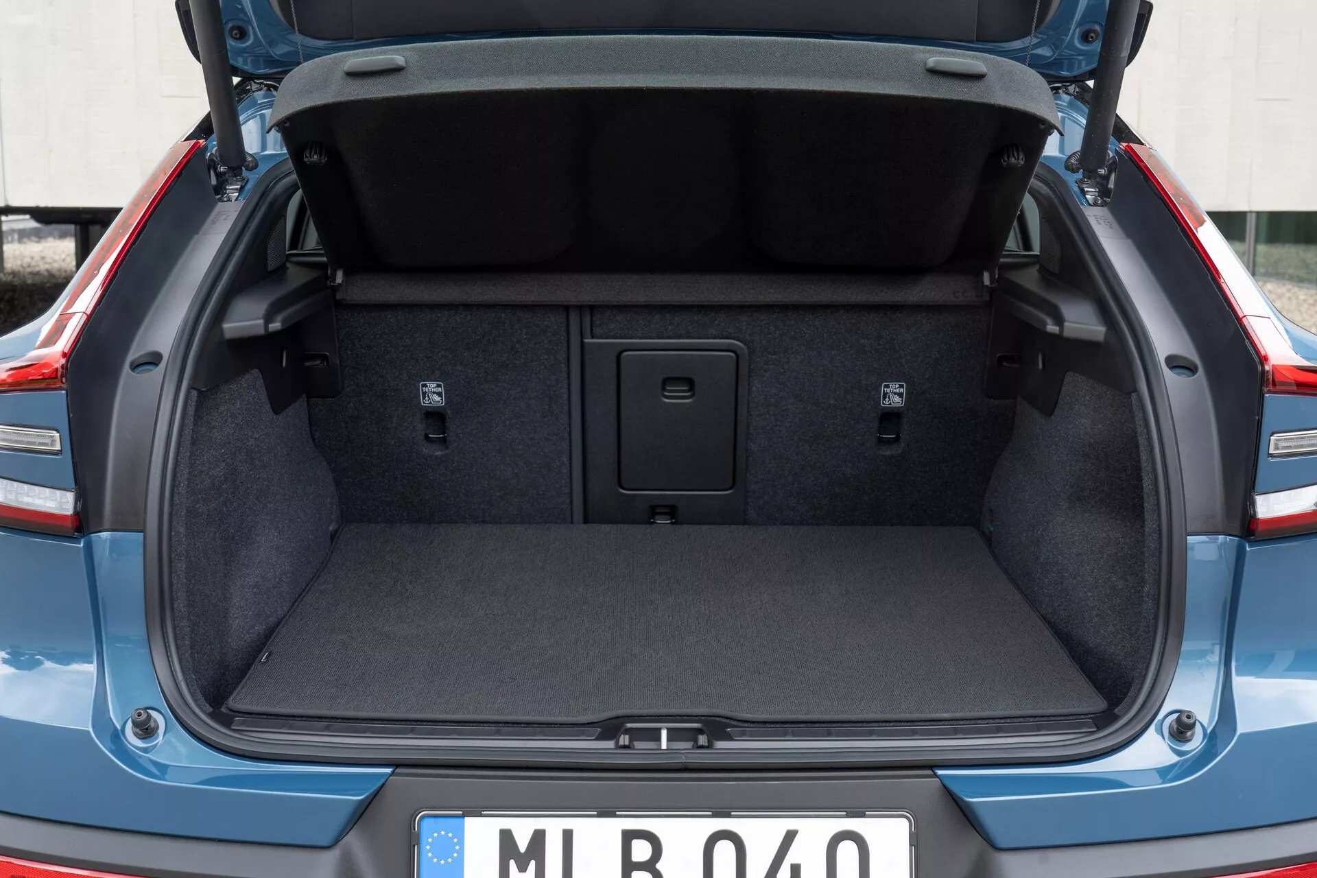 Spazio di carico posteriore di un'auto moderna, che mostra il bagagliaio della Volvo C40 2021 (1ª generazione) da una prospettiva posteriore, leggermente sotto il livello della vita. Il coperchio del bagagliaio è aperto, evidenziando l'imbottitura standard del rivestimento interno. Il bagagliaio è vuoto, mostrando l'interno tappezzato, le aree laterali incassate e una maniglia al centro del pavimento per l'accesso a un vano di stoccaggio. Non sono visibili ganci o punti di ancoraggio per il carico. La targa in stile europeo suggerisce un'origine europea.