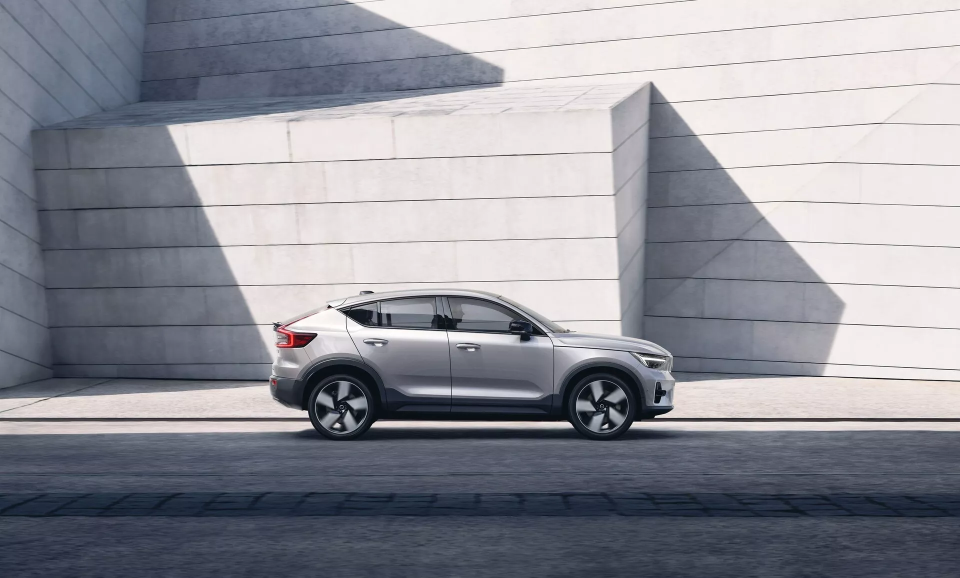 Immagine di un moderno SUV Volvo C40 2021 parcheggiato in parallelo, mostrando il profilo laterale sinistro dal faro anteriore sinistro alla luce posteriore sinistra. L'angolazione evidenzia la distanza dal suolo e la postura su una superficie pavimentata. La luce del sole crea ombre sulla finitura della vernice lucida. Il SUV presenta un design elegante con un tetto dolcemente inclinato, contorni distinti delle porte, vetri oscurati e specchietti laterali contemporanei. Le ruote in lega con un design multirazze e pneumatici a basso profilo indicano performance e lusso. Sono visibili la minigonna laterale, le modanature dei passaruota e una linea di carattere attraverso le maniglie delle porte, suggerendo robustezza. L'assenza di visibile scarico suggerisce una possibile propulsione elettrica o ibrida. Le strutture in cemento minimaliste sullo sfondo contrastano con le curve raffinate dell'auto, sottolineando il suo appeal urbano e di design.
