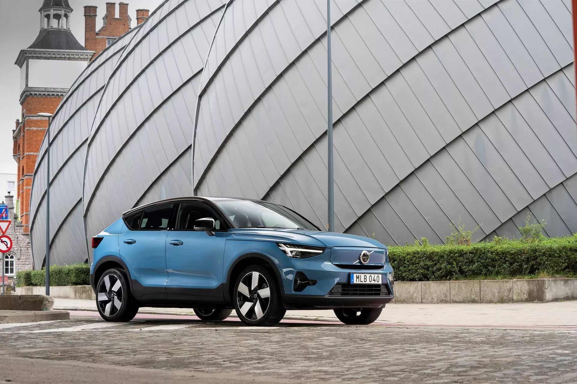 Un moderno SUV Volvo C40 2021 blu parcheggiato su un'area pavimentata urbana con una struttura angolare dall'aspetto metallico sullo sfondo. La ripresa a bassa angolazione dal lato anteriore sinistro mostra la griglia frontale del veicolo, i fari a LED, il profilo aerodinamico, i cerchi in lega a più raggi, gli specchietti laterali con indicatori di direzione integrati, la finitura della vernice opaca o satinata, i vetri oscurati, la targa anteriore e le linee scolpite del corpo.