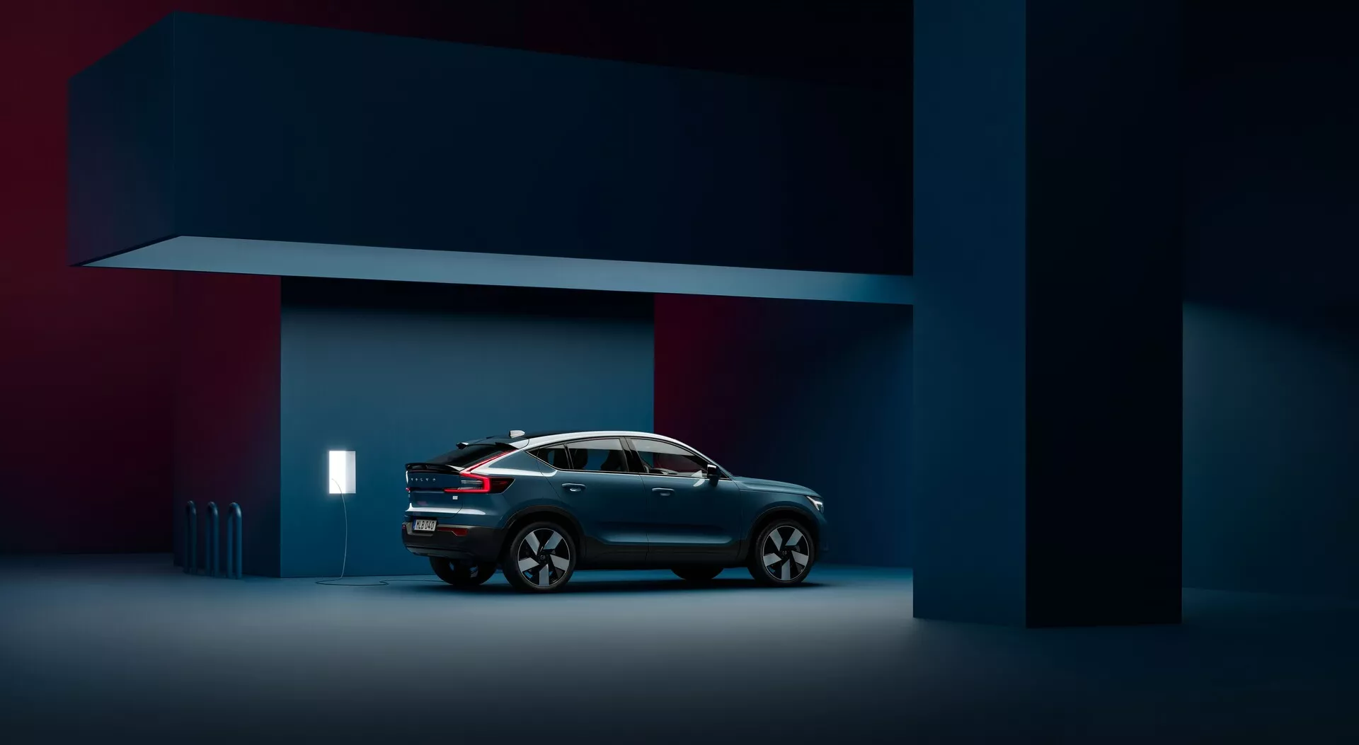 Vista di profilo laterale della Volvo C40 2021 di 1ª generazione in un ambiente studio, che mostra l'intera lunghezza del veicolo contro uno sfondo minimalista a due tonalità di blu navy scuro. La macchina, posizionata centralmente, è ripresa con un'illuminazione ambientale che evidenzia il suo design moderno e elegante crossover coupé-SUV, mettendo in risalto i suoi specchietti laterali aerodinamici, le ruote in lega uniche con raggi, le maniglie delle porte a filo e il sistema di illuminazione a LED. La vernice metallica interagisce con la morbida illuminazione dello studio, accentuando il gradiente di colore e le linee del veicolo.