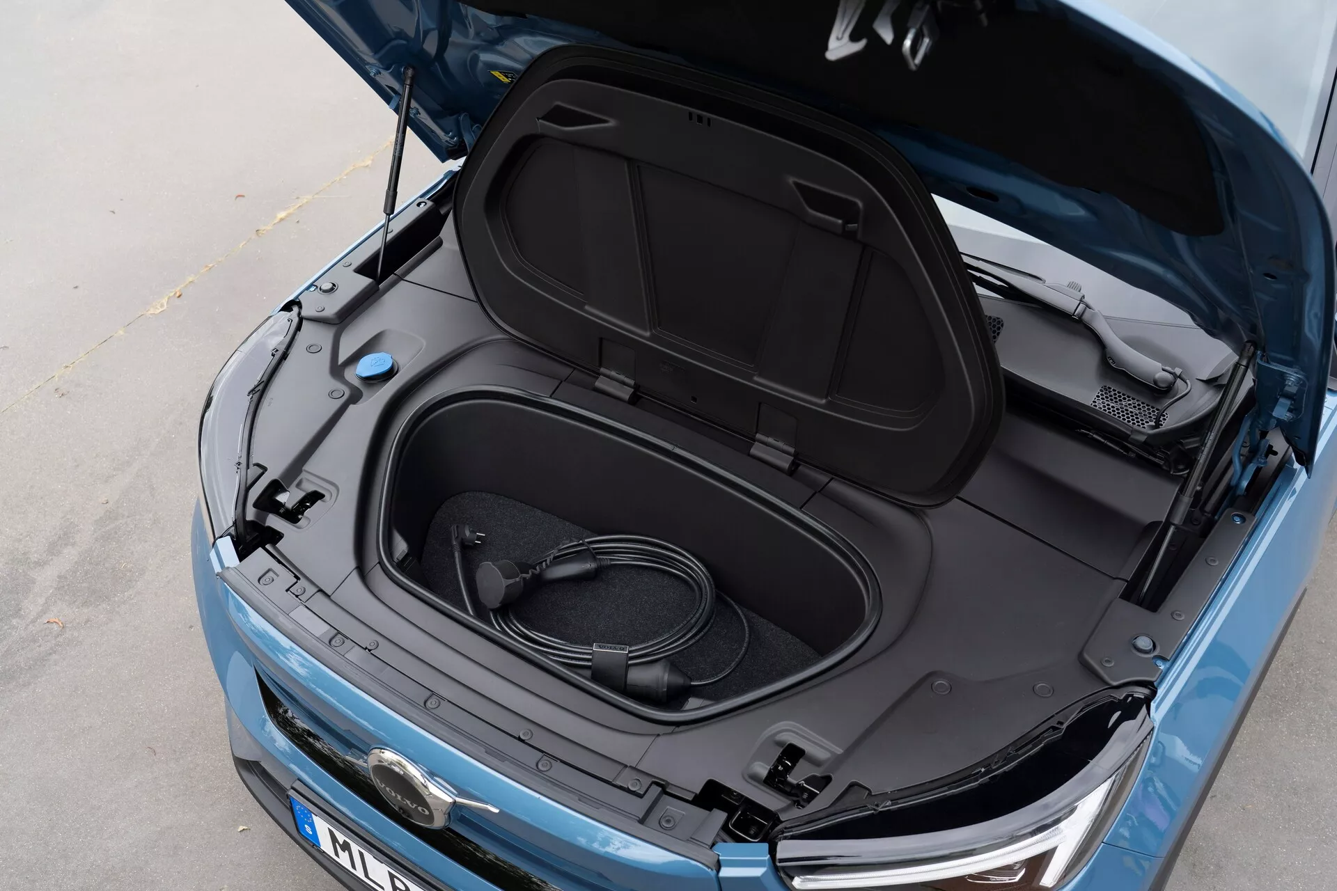 Vista ravvicinata dall'alto angolata della Volvo C40 2021 (1ª generazione) con il frunk aperto, che rivela spazio di stivaggio vuoto e un cavo di ricarica arrotolato, indicando che si tratta di un veicolo elettrico. Vernice blu metallizzato, interno del coperchio del frunk nero con adesivi informativi, vista parziale del logo del produttore, fari eleganti e griglia anteriore visibili, suggerendo un EV con tecnologia LED moderna ed estetica aerodinamica.