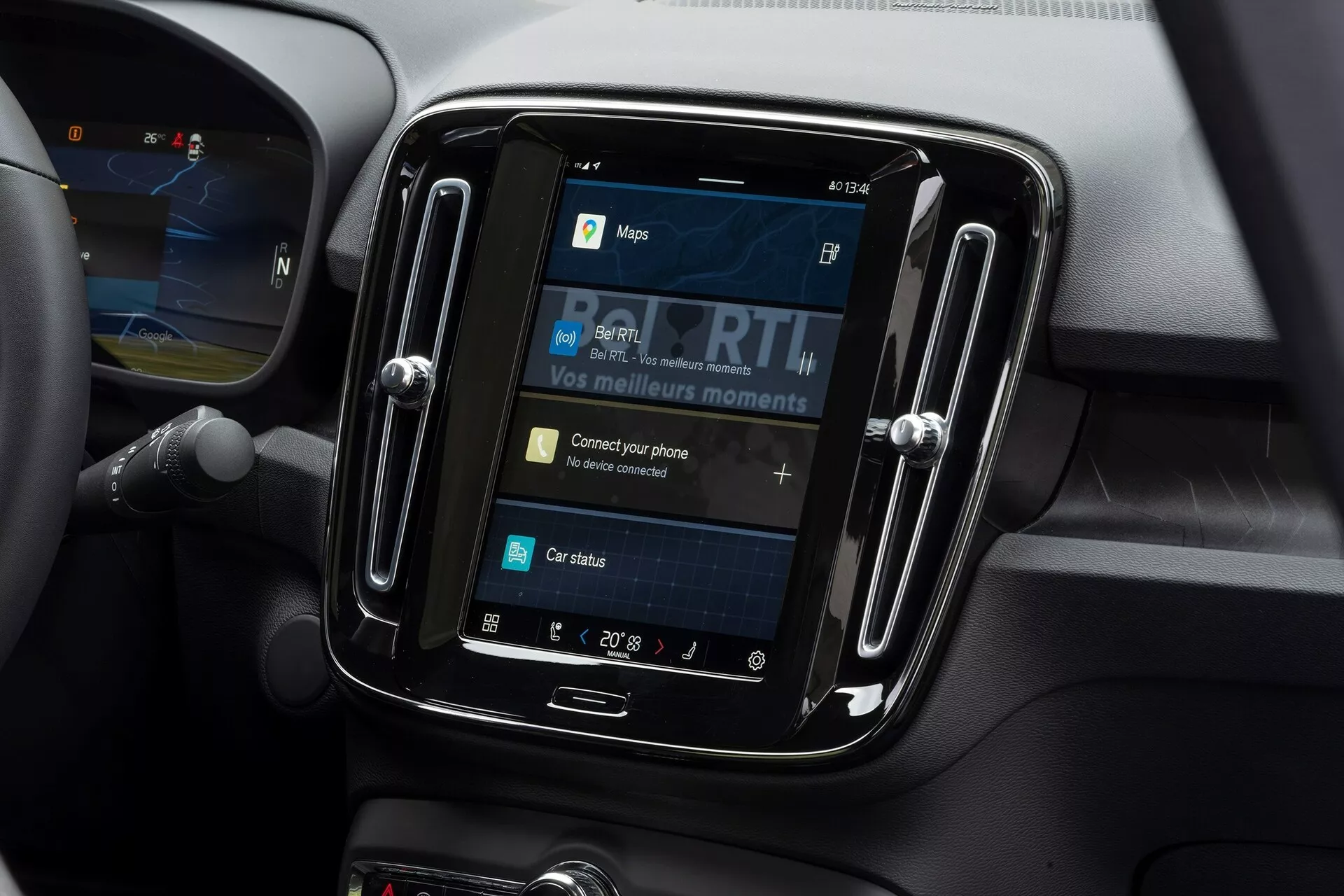 Vista ravvicinata del sistema di infotainment all'interno di una Volvo C40 2021. La ripresa, effettuata da un'angolazione leggermente rivolta verso il basso, si concentra sul display touchscreen incassato in una cornice nera lucida. Lo schermo mostra opzioni come mappe, media, connettività del telefono e stato dell'auto, con 'Connetti il telefono' come elemento interattivo. Intorno al sistema di infotainment ci sono materiali soft-touch e linee di contorno che indicano prese d'aria, suggerendo un interno auto moderno. L'impostazione della temperatura mostra 20° Celsius e parte del volante è visibile sul lato sinistro.