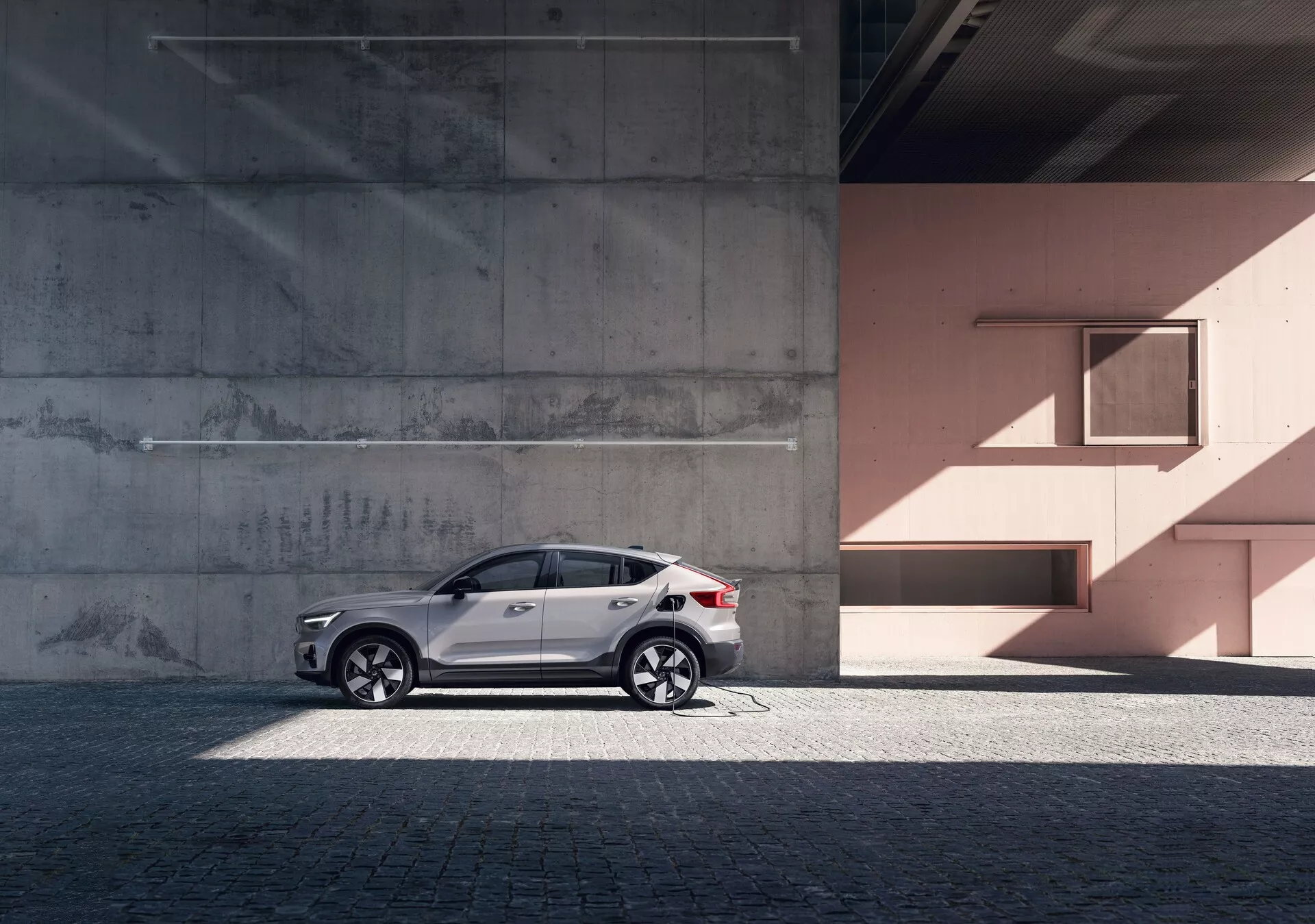 Profilo laterale del moderno SUV Volvo C40 2021 (1ª Generazione) contro uno sfondo urbano con pareti di cemento e color pesca. L'auto presenta una vernice bianca riflettente, eleganti cerchi in lega, finestrini oscurati, barre del tetto argentate e indicatori di direzione integrati negli specchietti retrovisori. La foto evidenzia le linee e le curve del design dell'auto alla luce naturale del giorno, aggiungendo un senso di lusso e sofisticazione.