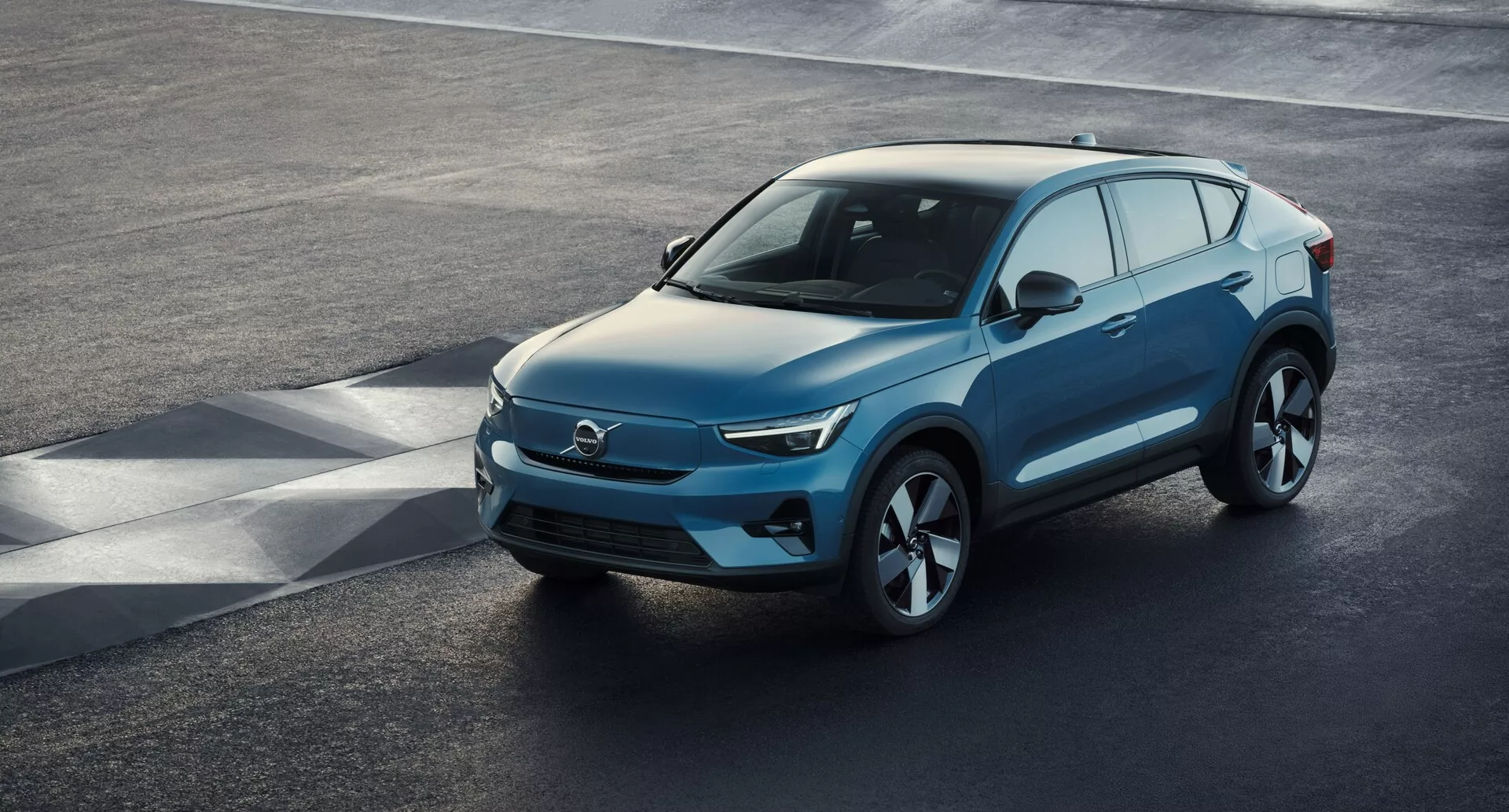Vista angolare elevata di un SUV moderno Volvo C40 del 2021 (1ª generazione) parcheggiato su una superficie di asfalto, vista di tre quarti anteriore dall'alto. La parte anteriore elegante, probabilmente caratteristica di un veicolo elettrico con prese d'aria minime, si combina con fari a LED nitidi, un cofano liscio in pendenza e un profilo laterale sagomato con grandi ruote in lega. Specchietti retrovisori laterali con segnale di svolta integrato, ampio spazio dei finestrini con possibile tinta di privacy e una linea del tetto dolcemente inclinata con barre sul tetto per utilità sono caratteristiche notevoli. La vista chiara cattura il design lussuoso del veicolo, l'aerodinamica avanzata e gli elementi tecnologici moderni.