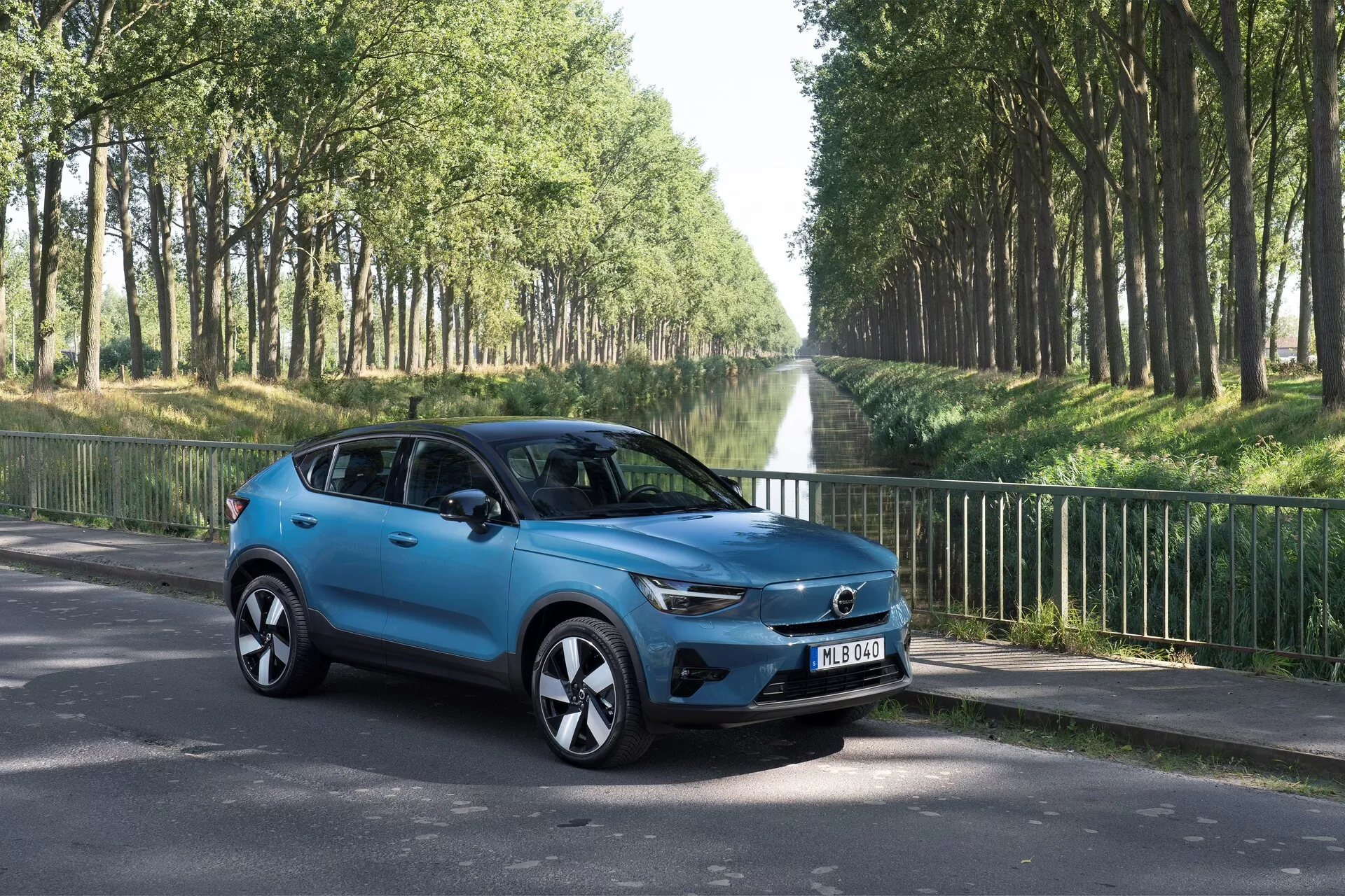 Volvo C40 2021 (Prima Generazione) SUV compatto in vista tre quarti da davanti a sinistra su strada asfaltata. Mostra elementi di design moderno come la griglia prominente con badge centrale, fari LED eleganti, cerchi in lega stilizzati, specchietti laterali con indicatori di direzione integrati, carrozzeria scolpita aerodinamicamente con una finitura di vernice blu brillante, vetri oscurati e quattro porte. Ambientato in un paesaggio esterno con alberi che costeggiano la strada, con focus sulle caratteristiche estetiche e tecnologiche del veicolo.