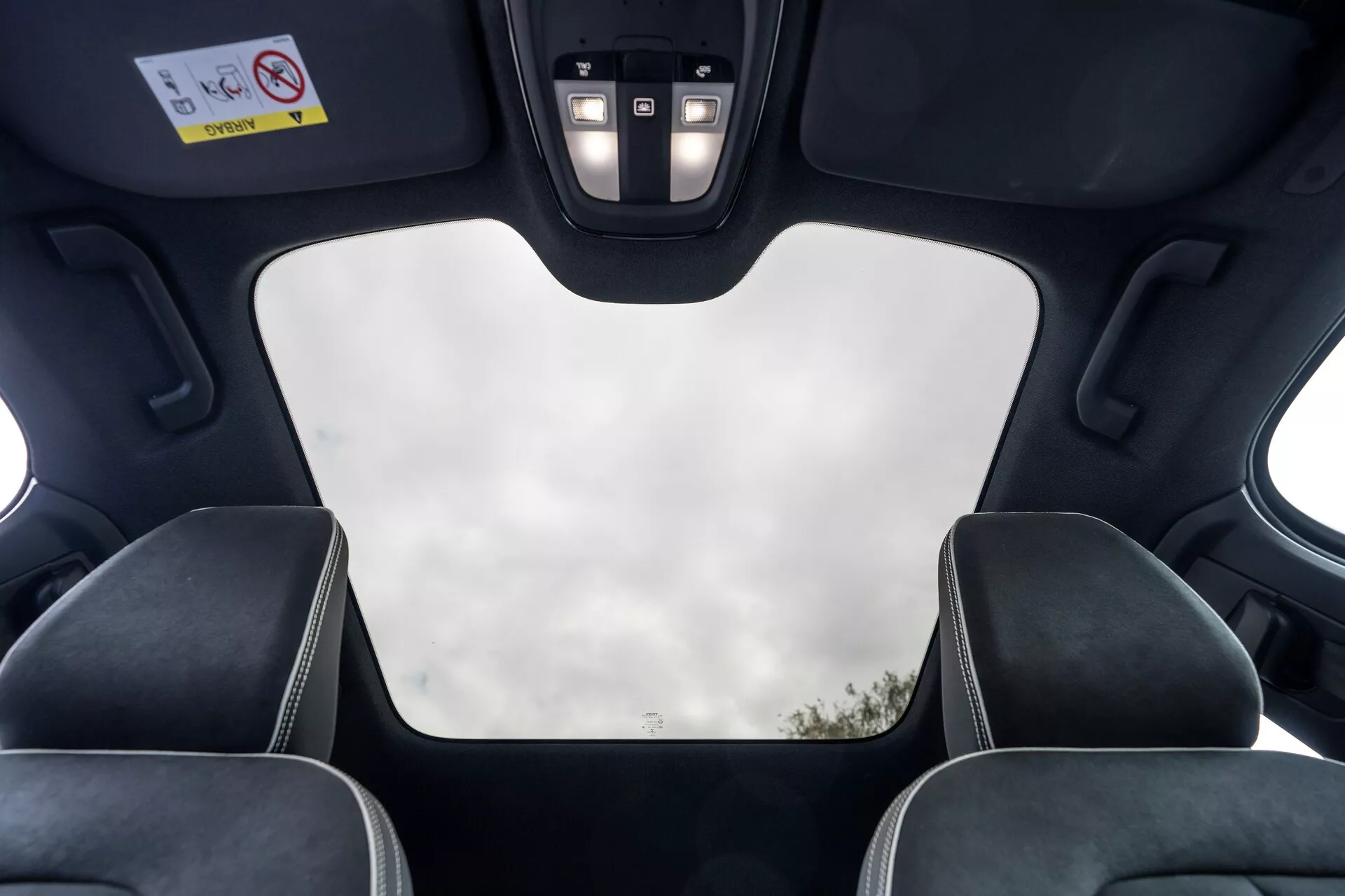 Vista interna della Volvo C40 2021 (1ª generazione) dalla prospettiva del passeggero posteriore, che mostra le caratteristiche del tetto come il tetto panoramico chiuso con vista chiara del cielo, rivestimento interno nero, intagli per il meccanismo del tetto, luci a LED e pannello di controllo con pulsanti per varie funzioni. L'immagine mette anche in risalto i poggiatesta dei sedili anteriori con cuciture a contrasto, enfatizzando il design moderno e lussuoso del veicolo e le sue caratteristiche di fascia alta.