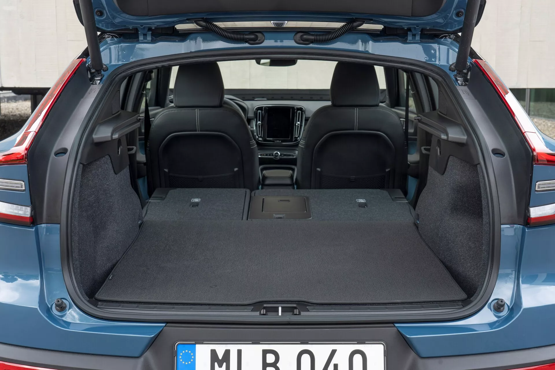 Interni posteriori della Volvo C40 2021 (1ª generazione) con il bagagliaio aperto, che mostra l'area di carico vuota con rivestimento in moquette resistente, superficie piatta e senza intrusioni visibili. È visibile la parte superiore dei sedili posteriori in tessuto scuro, con una visione parziale degli elementi della cabina anteriore come la console centrale e il sistema di infotainment. Il coperchio del bagagliaio è sollevato, rivelando i bordi rifiniti in plastica/composito.