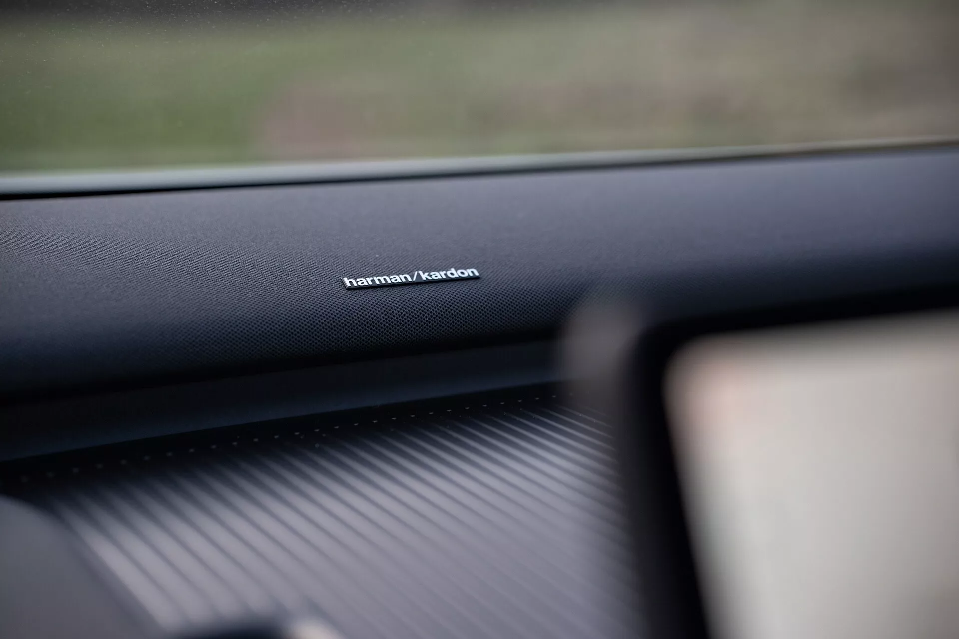 Scatto interno di un Volvo EX30 2024 con focus sul marchio del sistema audio Harman/Kardon sul cruscotto lato passeggero, evidenziando la qualità dei materiali di pregio e sottolineando la tecnologia avanzata del sistema sonoro.