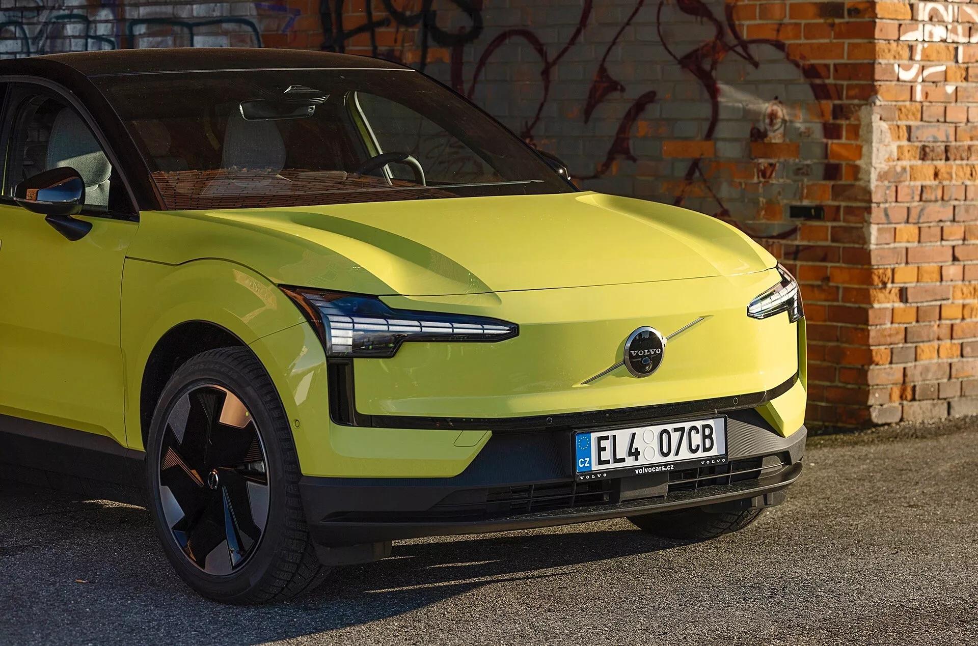 Auto elettrica Volvo EX30 verde lime del 2024 parcheggiata davanti a un muro di mattoni con graffiti, mostra design aerodinamico, fari LED moderni e assenza di griglia frontale