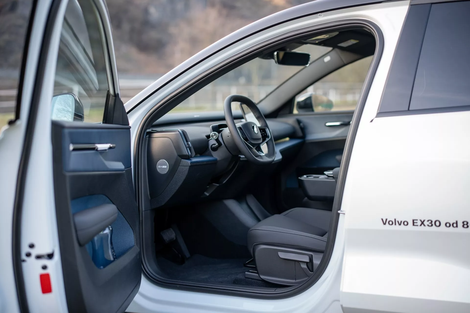 Vista interna di una Volvo EX30 2024 bianca, mostrando il sedile del conducente e l'area cockpit con la porta anteriore sinistra aperta dotata di comandi elettrici, e uno scorcio del volante e parte del cruscotto