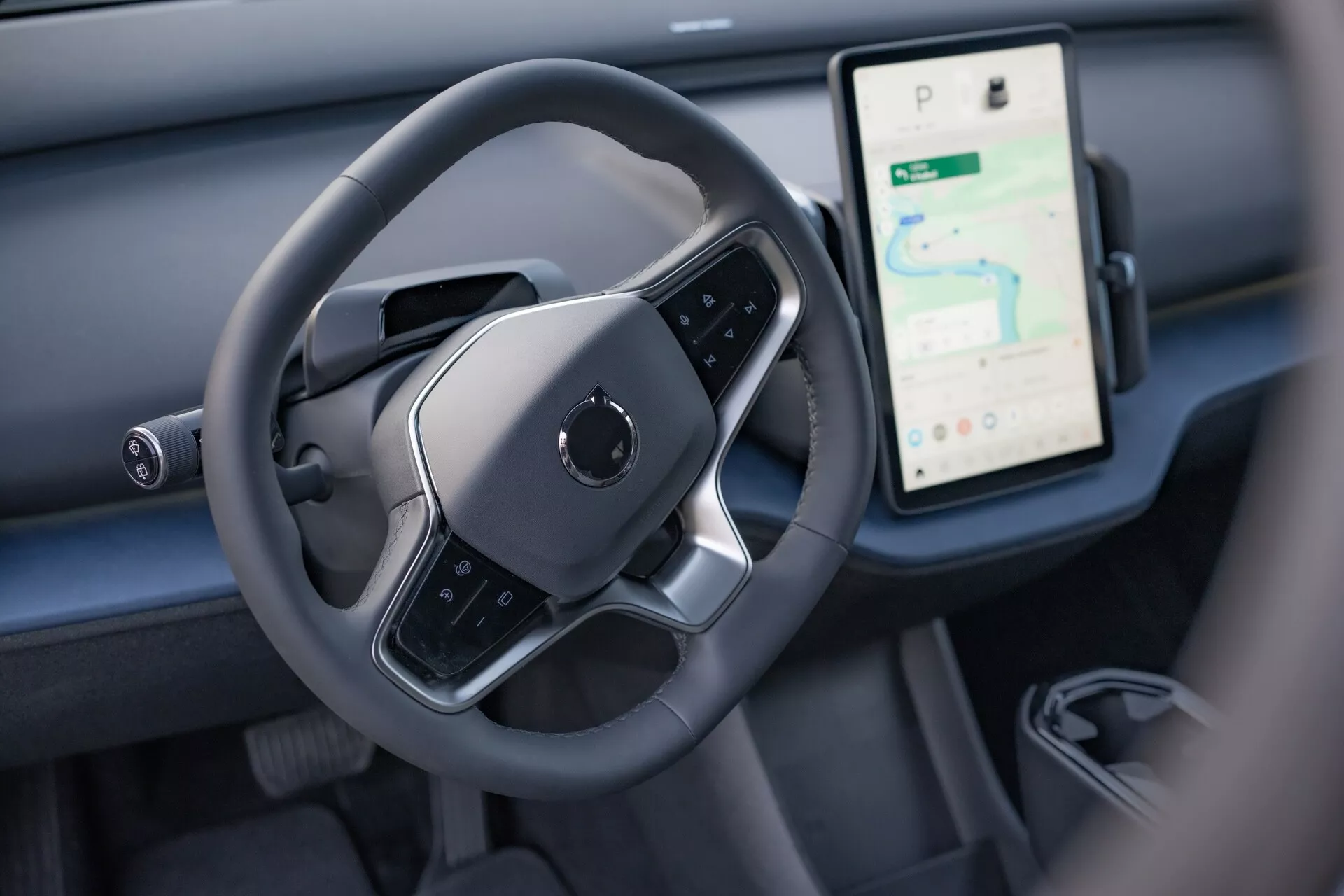 Vista interna del Volvo EX30 2024 che evidenzia l'area di controllo del conducente con un volante moderno a fondo piatto e un grande display touchscreen verticale per navigazione e media sul cruscotto