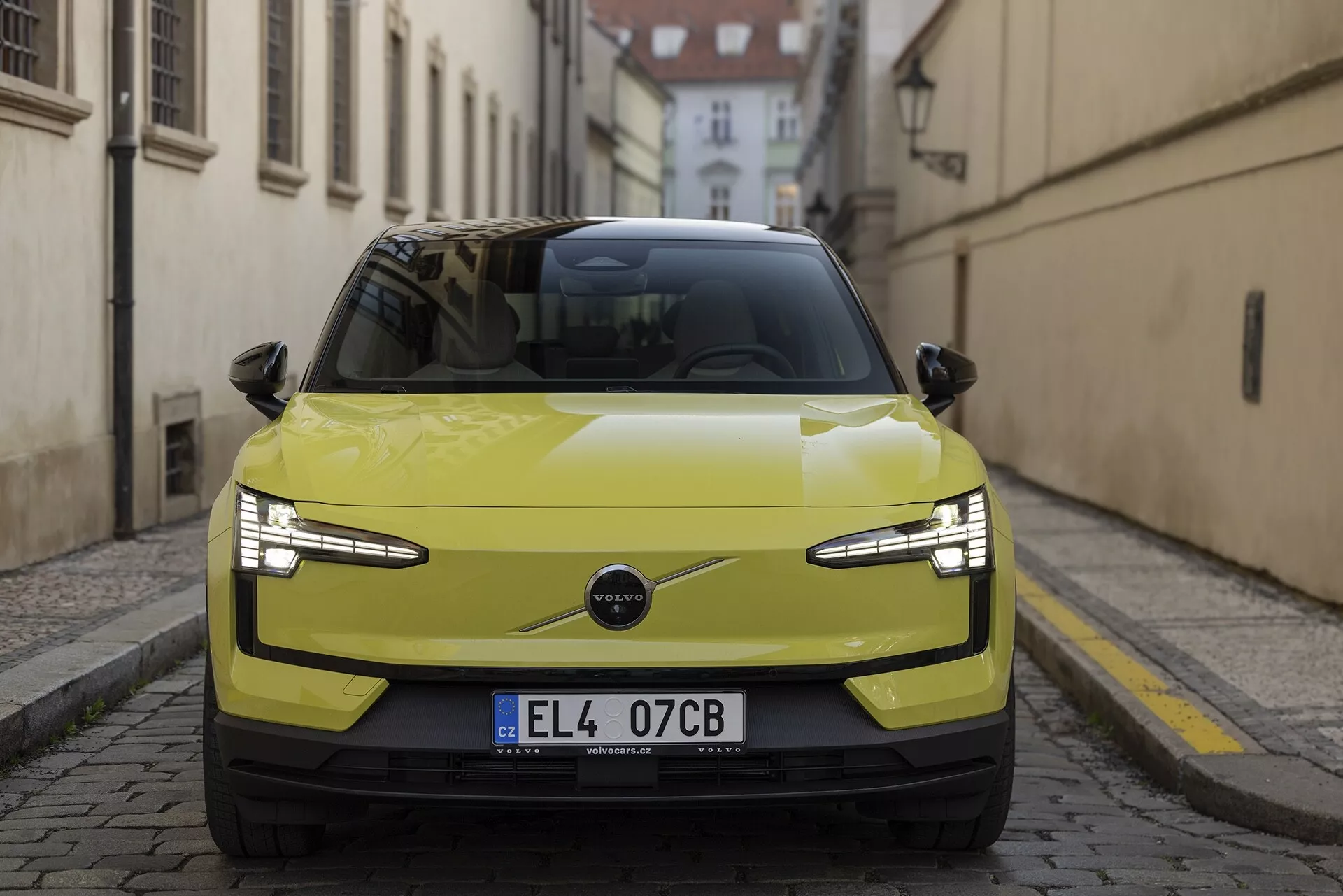 Vista frontale della Volvo EX30 2024 in giallo lime, con in evidenza i suoi fari LED eleganti, la griglia minimalista indicativa di un powertrain elettrico e caratteristiche tecnologiche avanzate come sensori di parcheggio e possibile ADAS, su uno sfondo di strada europea storica