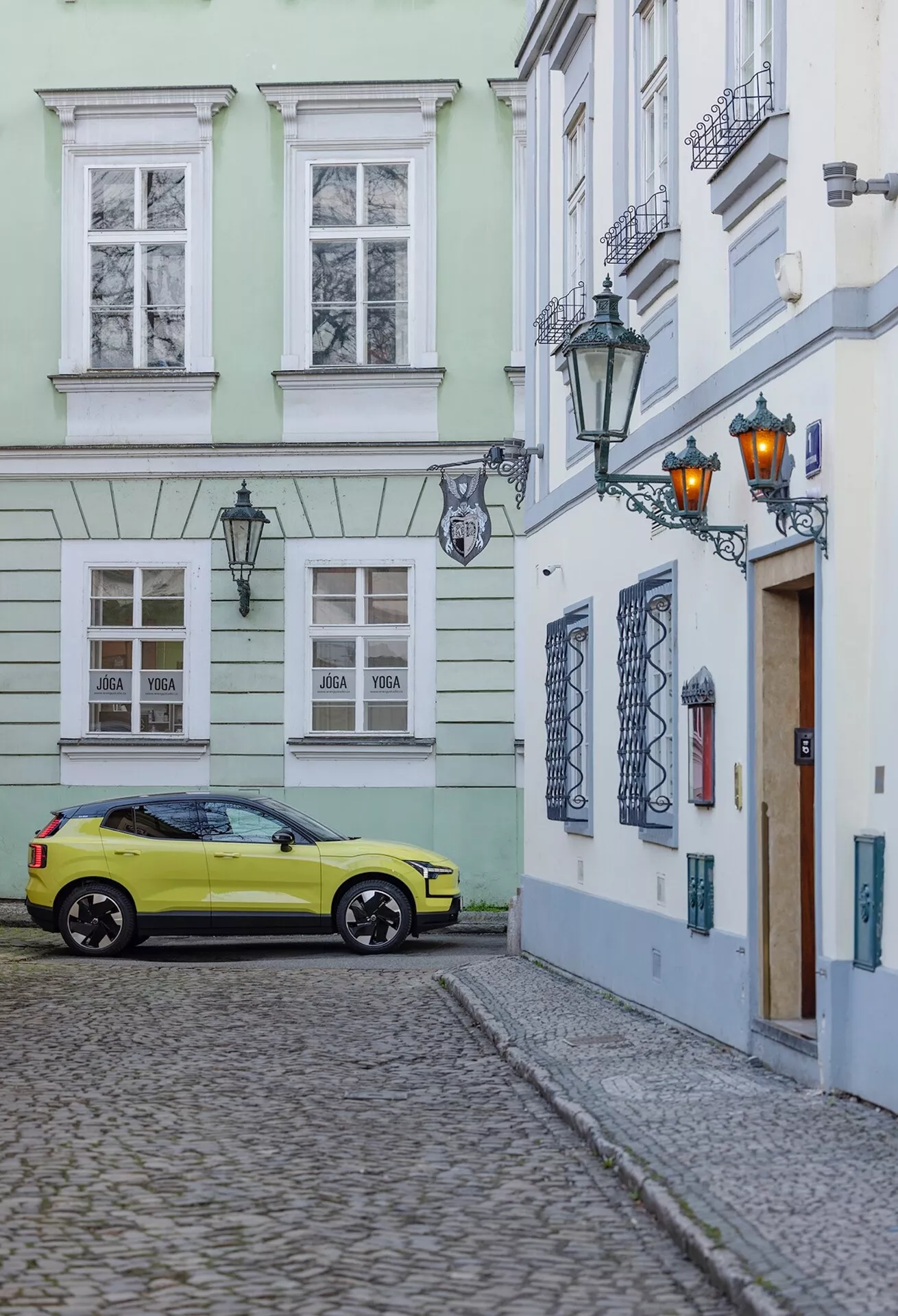 Volvo EX30 2024 parcheggiata su una strada lastricata in un contesto storico, ripresa da un angolo posteriore di tre quarti che mostra il lato destro e posteriore della macchina inclusi le ruote, i fanali posteriori e dettagli parziali del tetto in colore giallo acceso