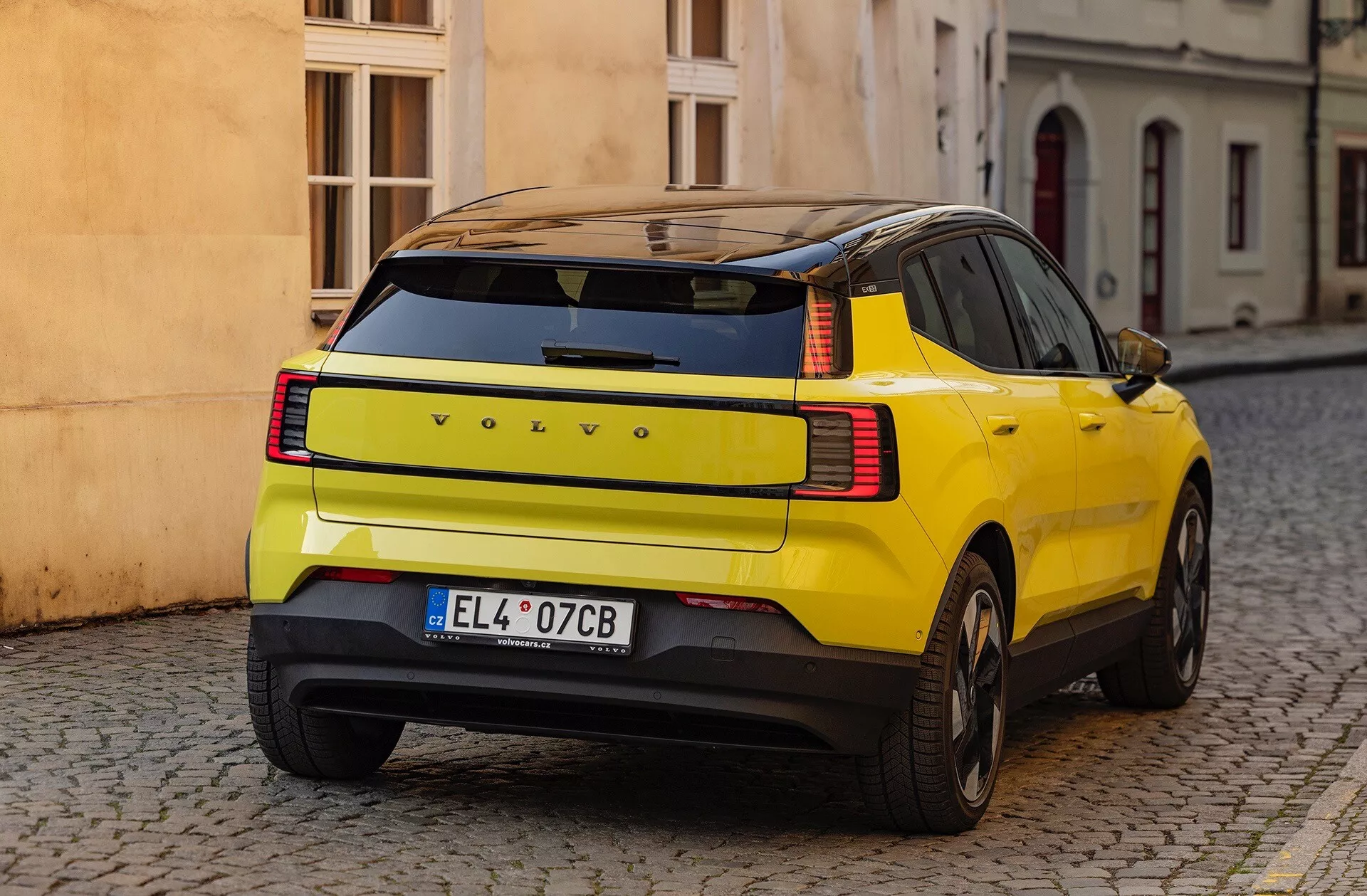 SUV giallo Volvo EX30 2024 mostra la sua distinta curvatura del pannello posteriore, luci posteriori LED e caratteristiche di sicurezza, parcheggiato su una strada in pietra da una vista posteriore di tre quarti