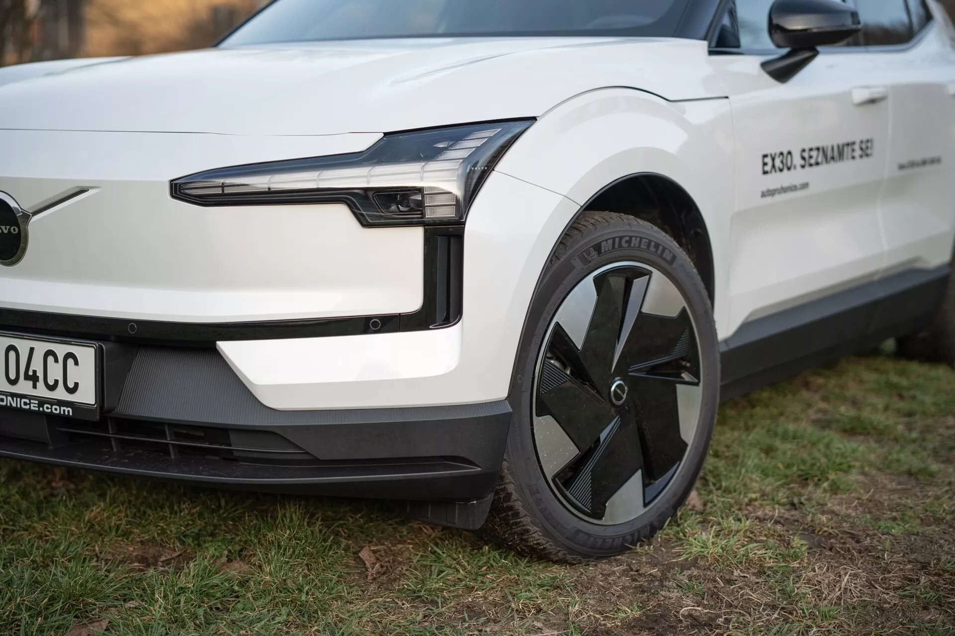 Vista ravvicinata del veicolo elettrico Volvo EX30 2024 da una prospettiva a tre quarti, evidenziando il suo faro LED, design aerodinamico della ruota e paraurti anteriore minimalista senza griglia, in bianco con dettagli neri.