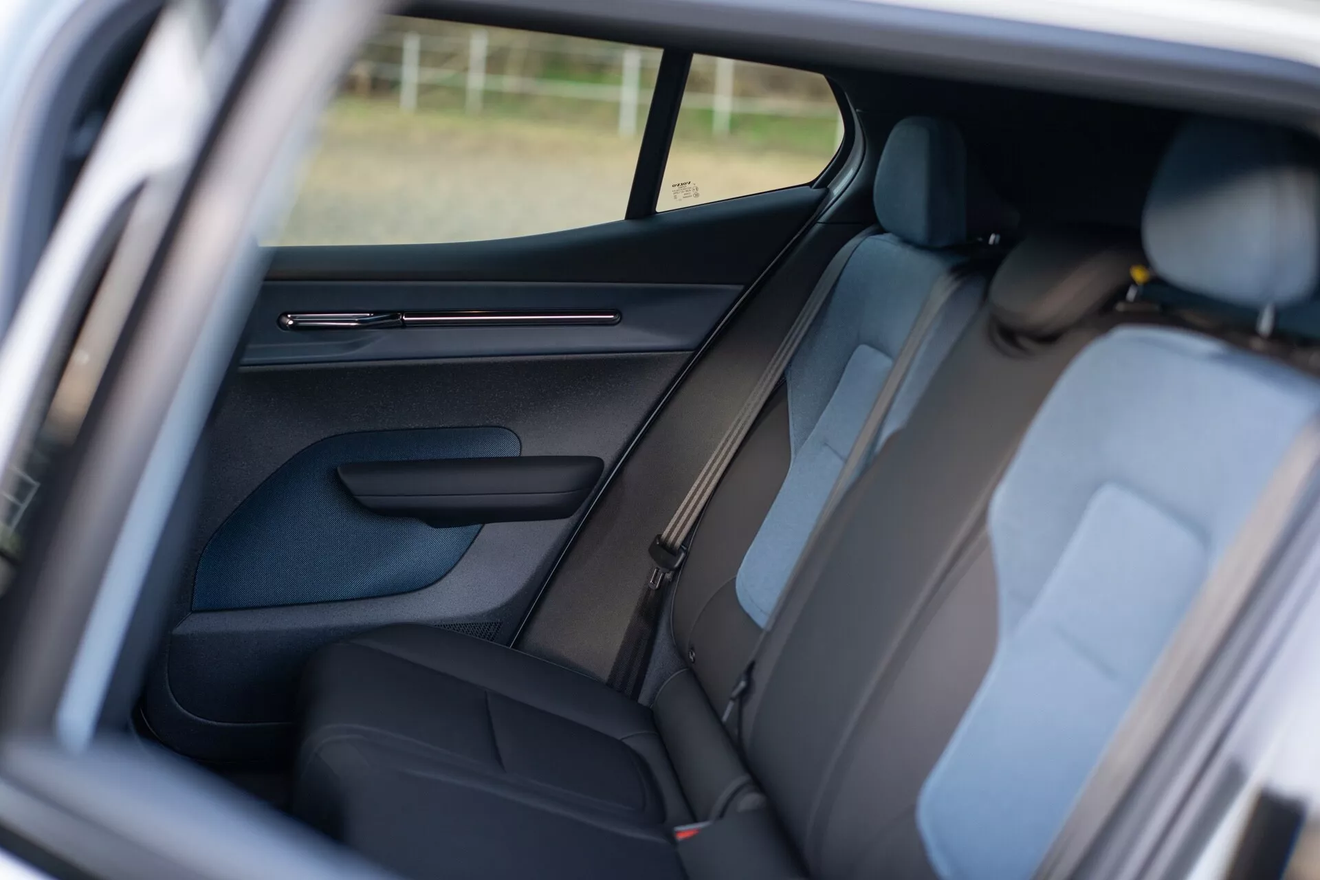 Interni Volvo EX30 2024 con vista sull'area passeggeri posteriore attraverso la porta anteriore aperta del passeggero, caratterizzati da tappezzeria scura, poggiatesta visibili, pannello porta dettagliato con comandi, design del montante B, cinture di sicurezza per i sedili posteriori, finestra laterale parzialmente visibile che migliora l'ambiente interno, e focus sulla disposizione dei sedili per il comfort e la sicurezza dei passeggeri.