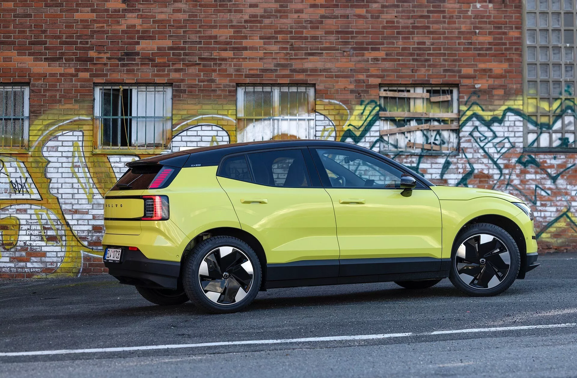 Immagine laterale di un SUV Volvo EX30 verde lime del 2024 parcheggiato accanto al bordo del marciapiede su strada urbana con arte graffito sullo sfondo, che mostra i suoi cerchi in lega nera sportivi, luci posteriori LED e luce stop alta montata.