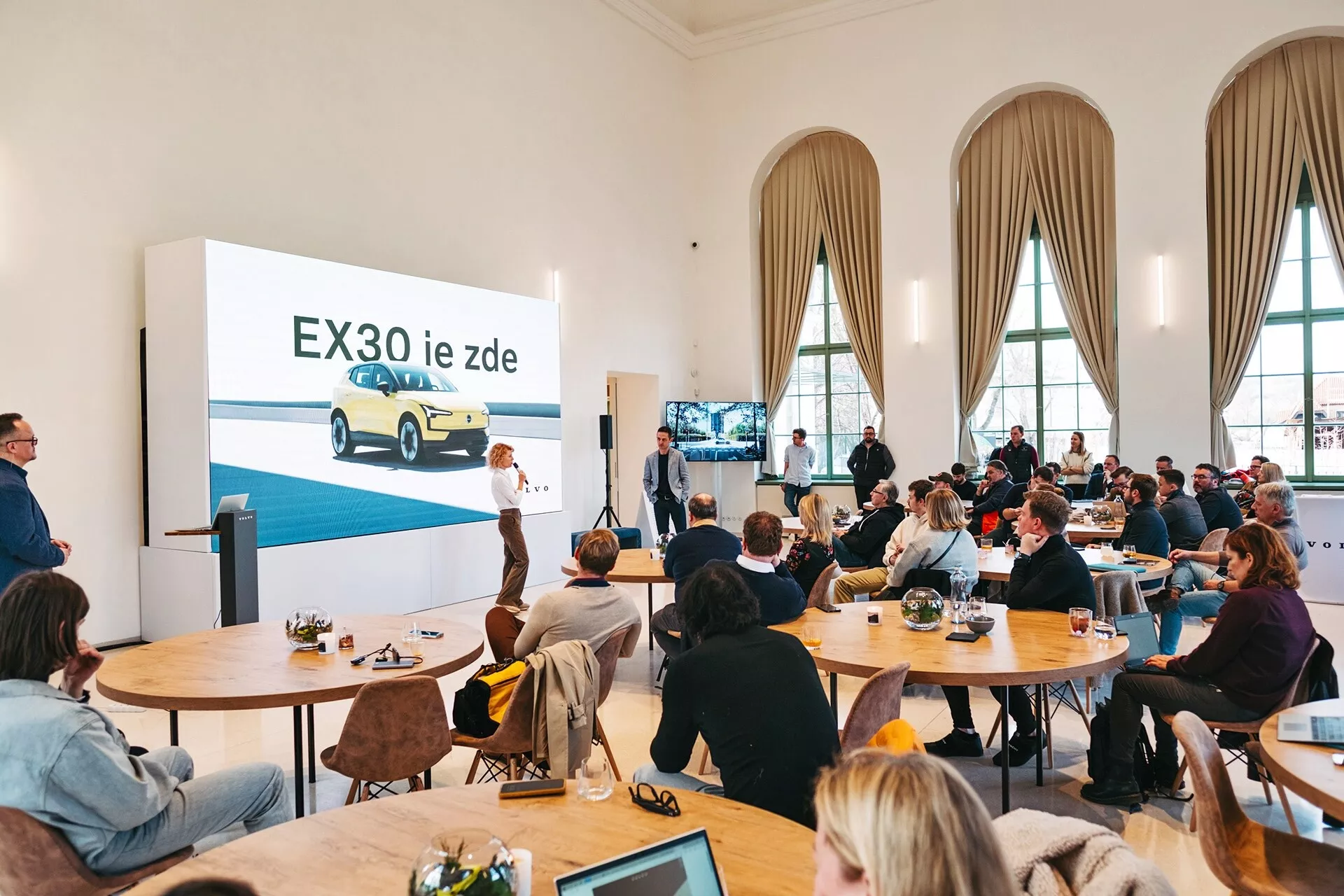 Presentazione della vettura elettrica Volvo EX30 2024 in una sala spaziosa con soffitti alti e grandi finestre ad arco, con un EX30 giallo visto da un angolo frontale di tre quarti su un grande schermo