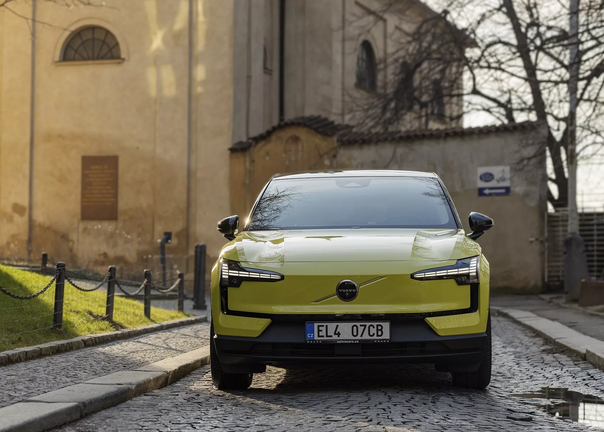 Automobile elettrica moderna Volvo EX30 del 2024 in colore chartreuse, parcheggiata su una strada lastricata, vista da una prospettiva frontale-destra con fari LED e design aerodinamico del cofano evidenziato contro uno sfondo storico europeo.