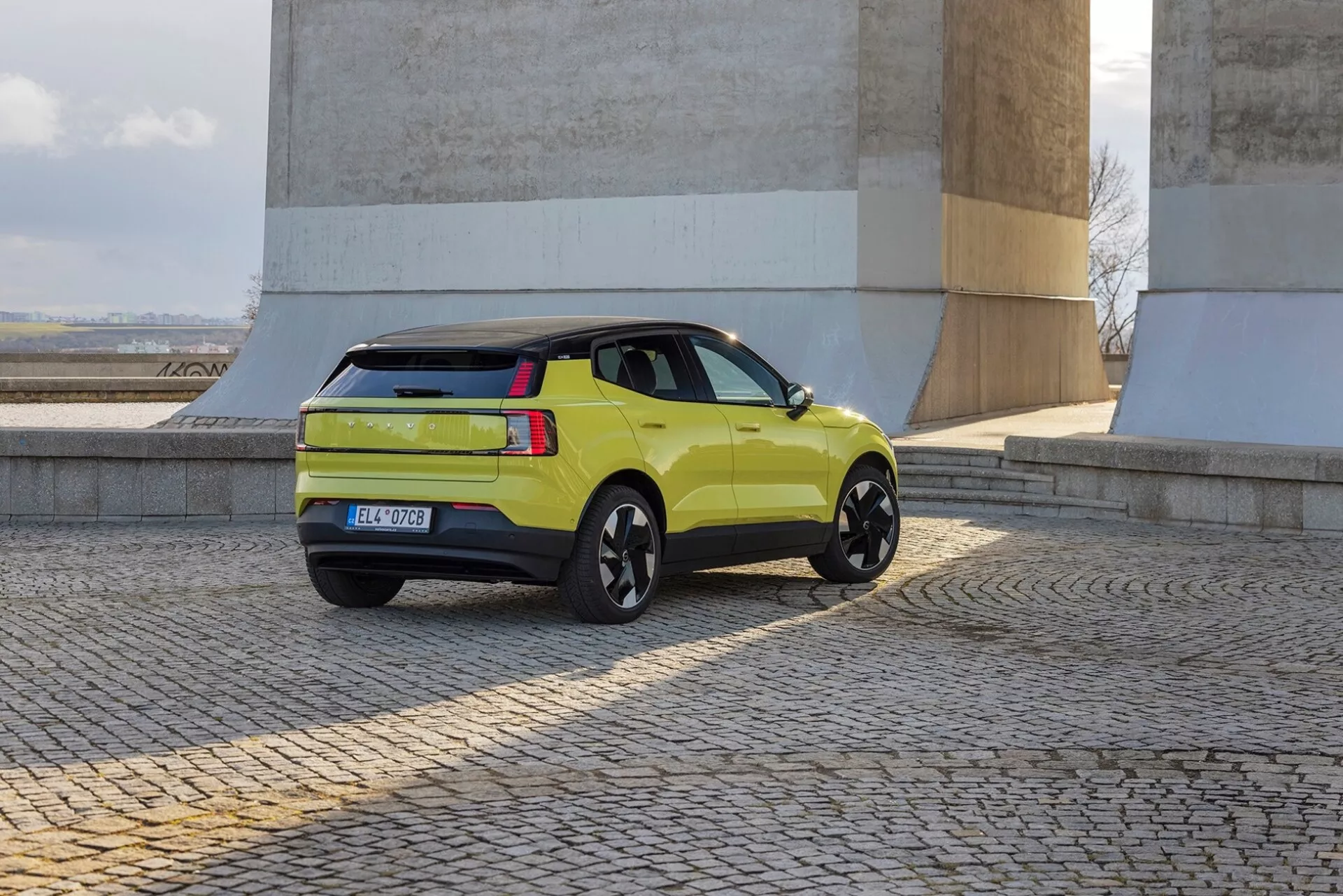 SUV verde lime Volvo EX30 2024 in veduta posteriore a tre quarti che mostra profilo laterale, luci posteriori, portellone e cerchi in lega neri vicino a muri in pietra su pavimento in ciottoli