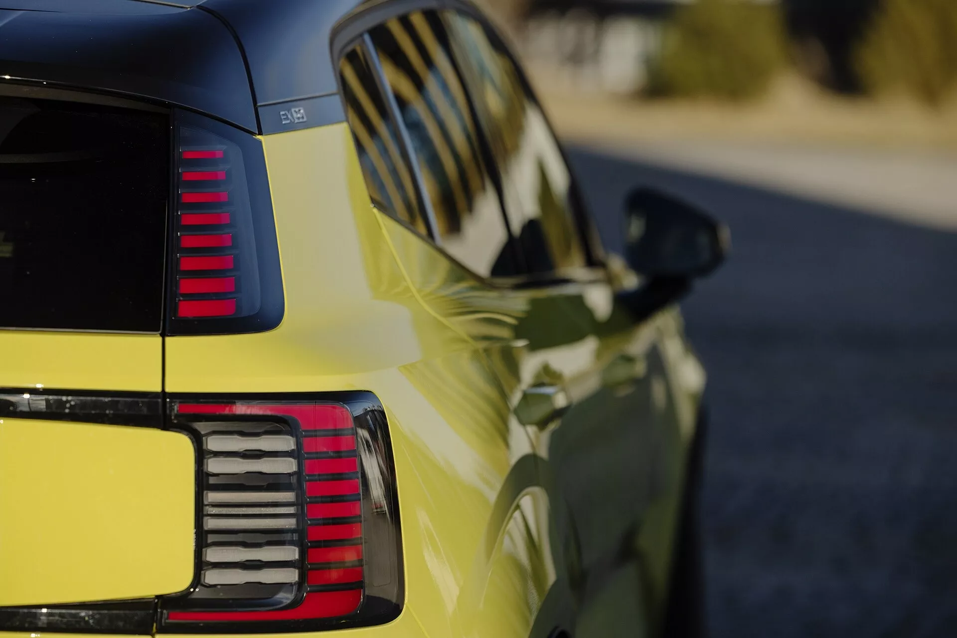 Vista ravvicinata del quarto posteriore di una Volvo EX30 2024 in giallo vibrante, mostrando la finitura lucida, i fanali posteriori LED segmentati, le linee di design aerodinamico e i segnali di svolta integrati nello specchietto laterale
