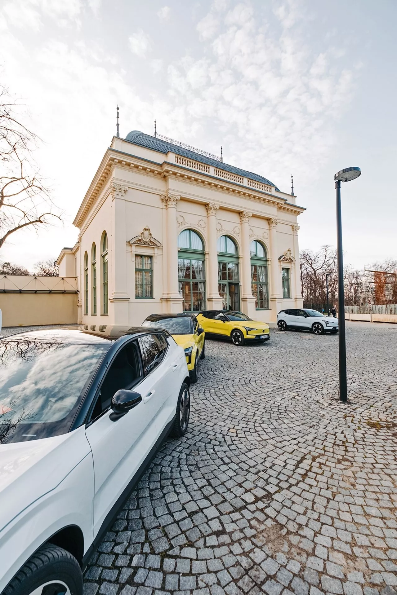 Scena stradale con tre auto ed un edificio elegante sullo sfondo, ripresa da una prospettiva grandangolare che evidenzia una berlina contemporanea bianca, una sportiva giallo vivace e una berlina o hatchback bianca sotto la luce del tardo pomeriggio