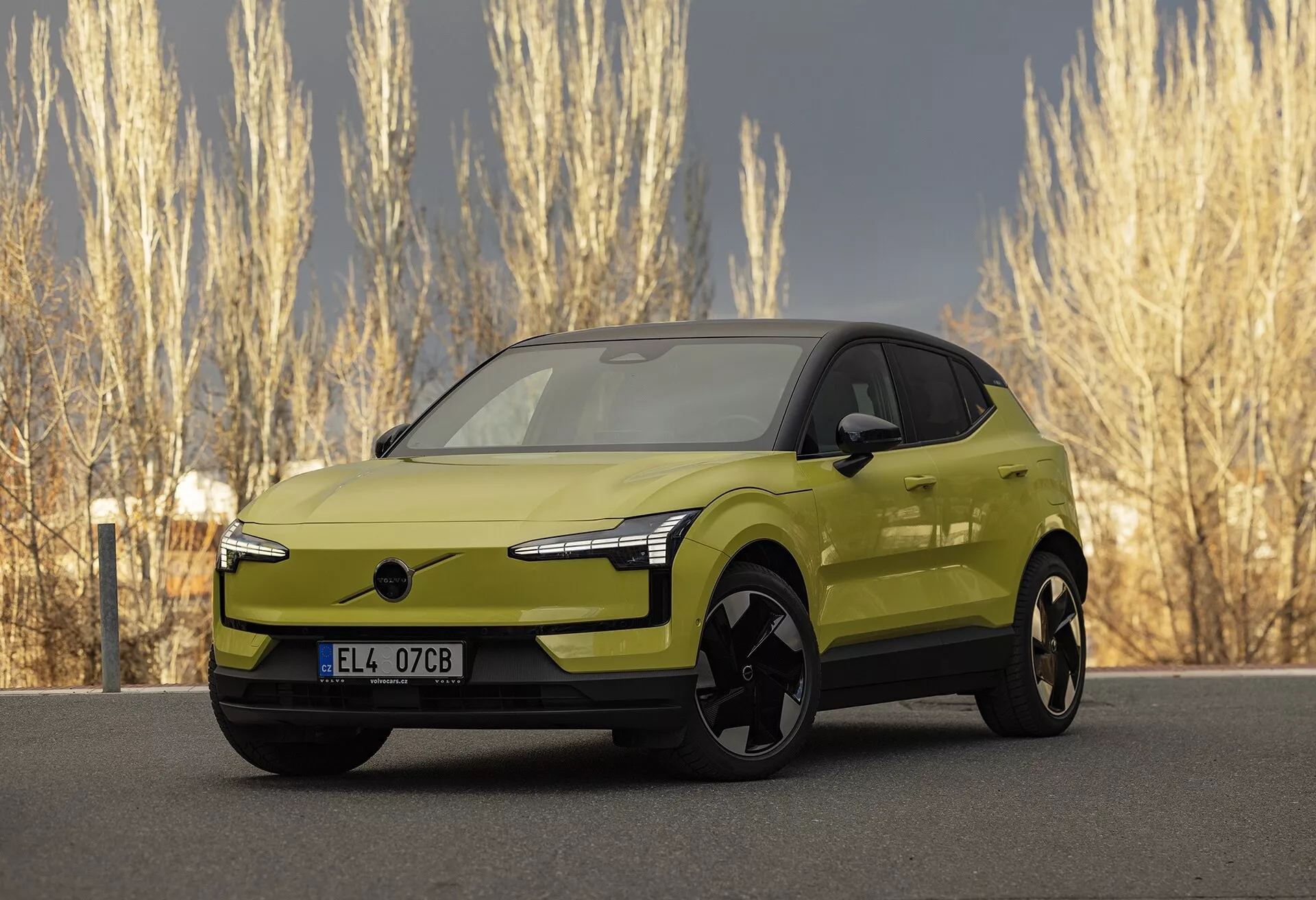 Moderna auto elettrica Volvo EX30 2024 di colore verde lime in vista frontale tre quarti che evidenzia i suoi fari LED eleganti, l'assenza di griglia tradizionale per migliorare l'aerodinamica, grandi ruote in lega nera, specchietti esterni color carrozzeria con potenziale tecnologia integrata, profilo laterale liscio e aerodinamico con un aspetto simile a un coupé, e finitura metallica vivace, parcheggiata in un ambiente naturale