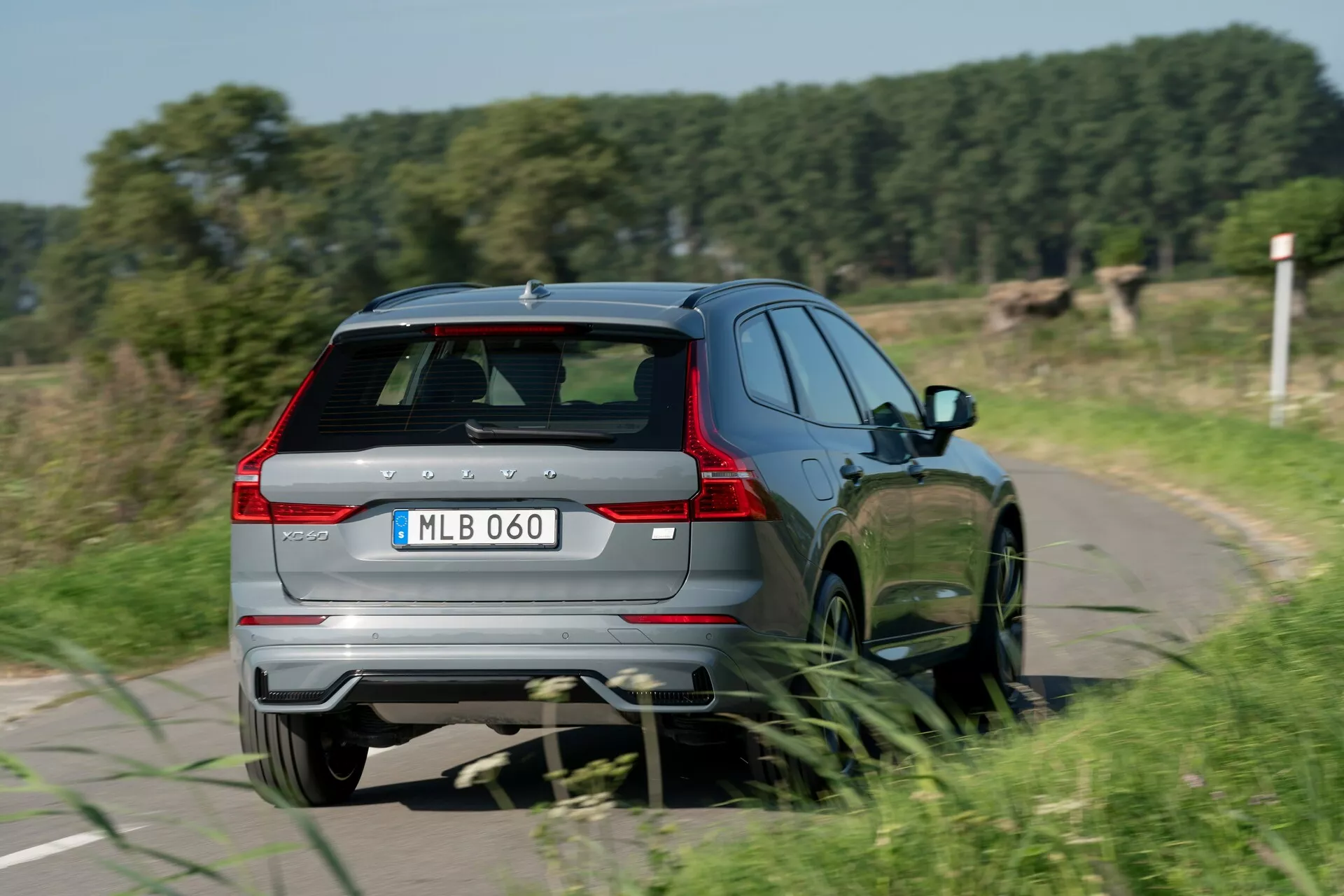 Vista posteriore tre quarti di una Volvo XC60 2021 (seconda generazione facelift) in movimento, che mostra gli elementi di design della parte posteriore e del lato destro, tra cui il portellone con tergicristallo posteriore, spoiler e terza luce di stop, paraurti posteriore con riflettori integrati e sistema di scarico doppio, luci posteriori a LED allungate, profilo laterale destro pulito con porta passeggero posteriore e cerchi in lega multiraggi, e badge del modello. L'auto sta guidando su una stretta strada di campagna, catturando una sensazione di movimento con un leggero sfocatura delle ruote.