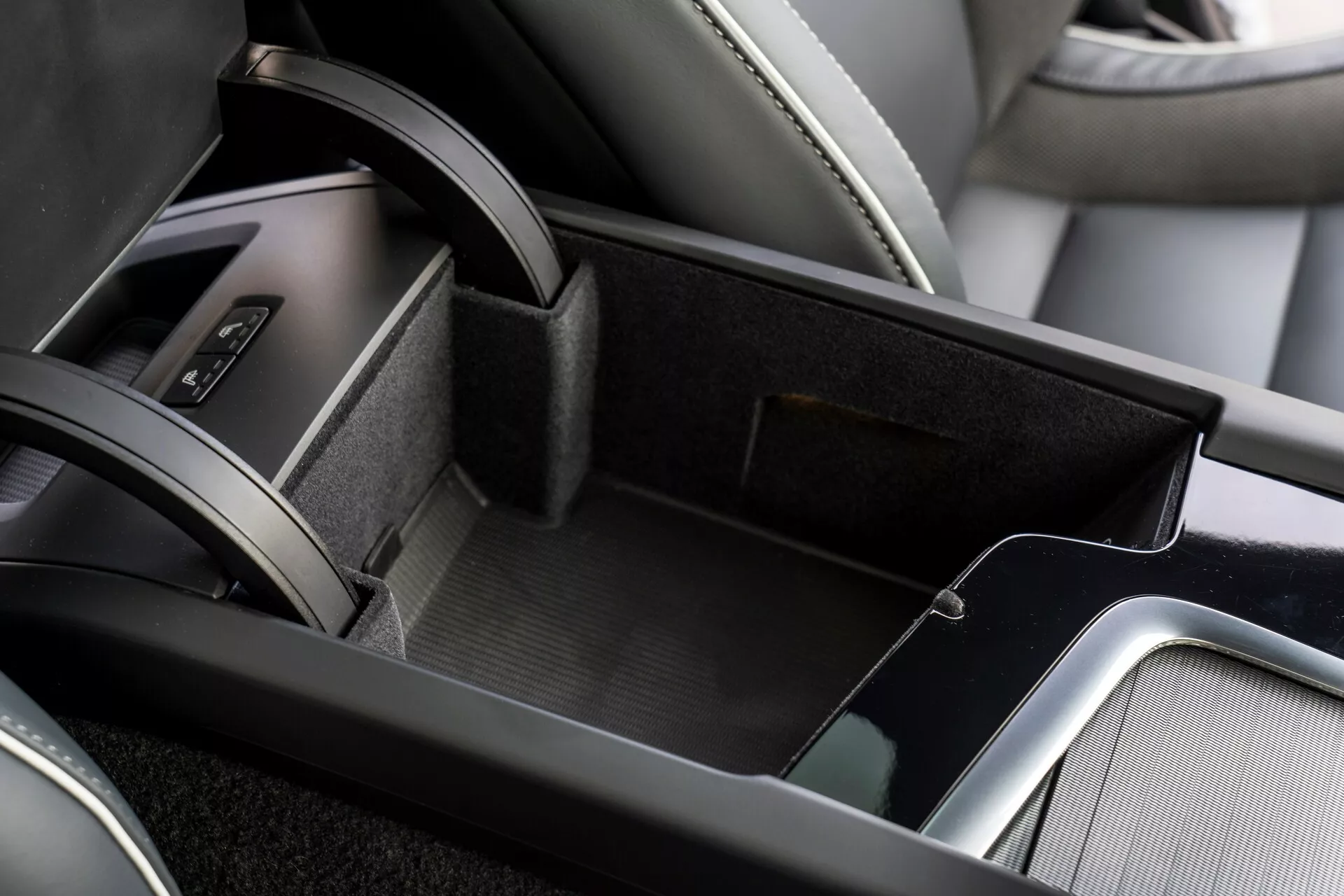 Vista ravvicinata dell'area console centrale nell'interno della Volvo XC60 2021, che mostra il vano di stivaggio aperto foderato in materiale simile al feltro, il vano portaoggetti a doppio livello con un vassoio superiore più piccolo, il bracciolo rivestito in pelle e le finiture metalliche che aggiungono raffinatezza. Parte dell'area della leva del cambio con rivestimento in pelle è visibile al bordo dell'immagine.