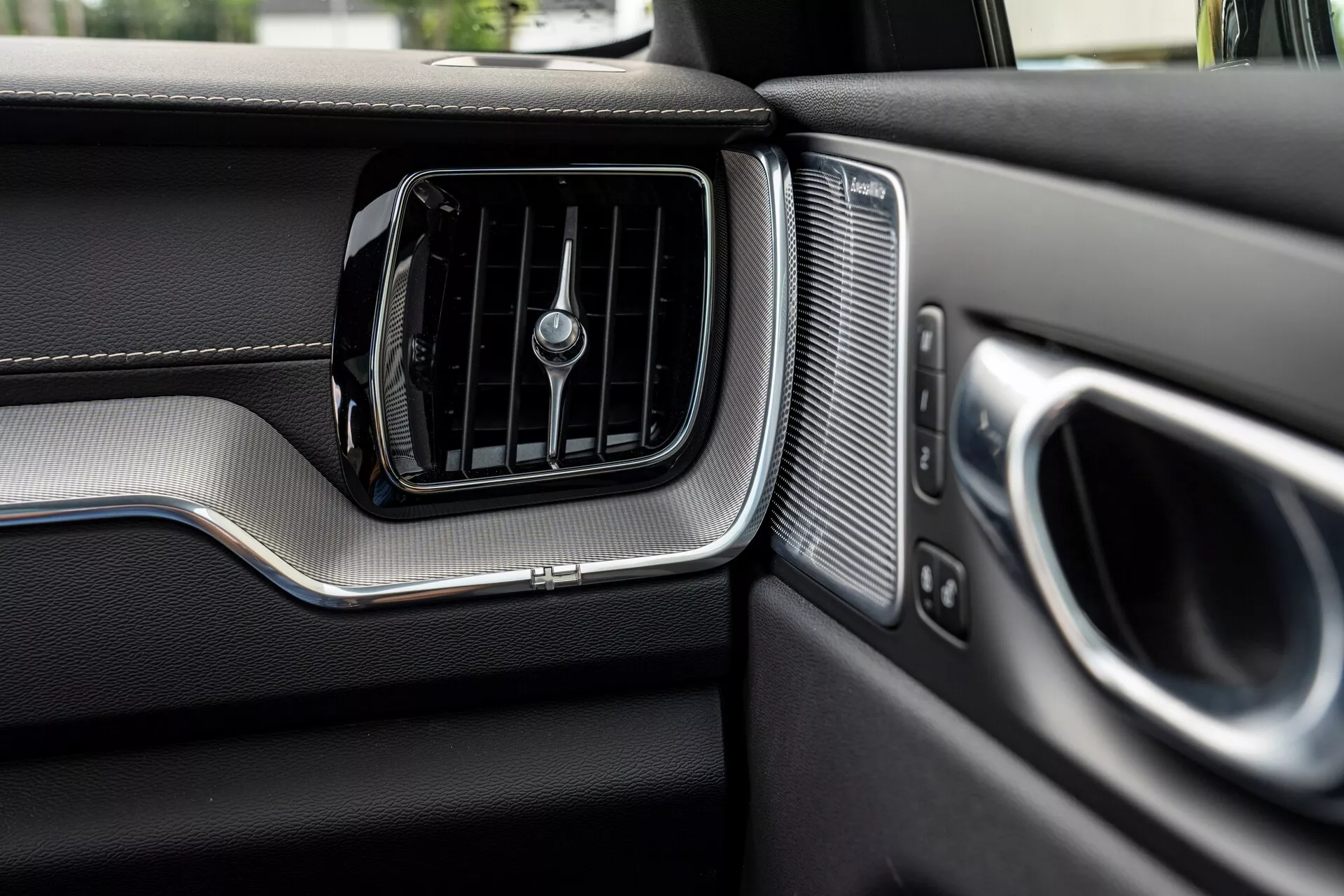 Vista ravvicinata del cruscotto lato passeggero in una Volvo XC60 2021 (2a generazione restyling). L'immagine mostra in primo piano la presa d'aria condizionata con lamelle orizzontali e una manopola centrale rotante, bordata da un accento metallico spazzolato. A destra della presa d'aria, c'è una griglia altoparlante con perforazioni uniformi, accanto al bracciolo della portiera e alla maniglia parzialmente visibili, anch'essi con accenti metallici. Il cruscotto presenta materiali simili alla pelle nella parte superiore e una zona inferiore di diversa trama separata da una striscia metallica, evidenziando il design premium e la qualità artigianale del veicolo.