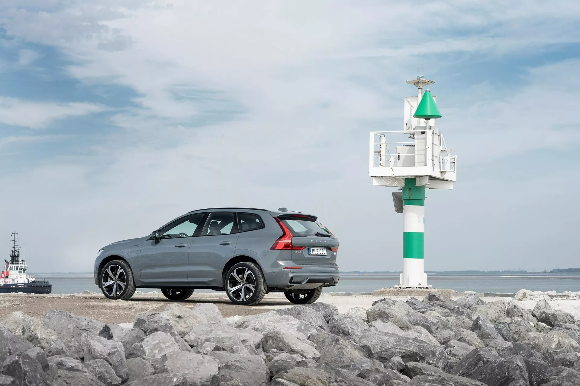 Fotografia del modello Volvo XC60 2021 di seconda generazione con restyling, catturata da un angolo posteriore di tre quarti su una superficie rocciosa, con uno sfondo marittimo. L'auto grigio scuro con finitura metallica presenta una linea del tetto slanciata e sportiva, cerchi in lega a più razze, sensori di parcheggio posteriori e doppio scarico. A destra dell'auto è visibile un piccolo faro o segnale luminoso, con un rimorchiatore o barca pilota nell'acqua, migliorando l'atmosfera costiera.