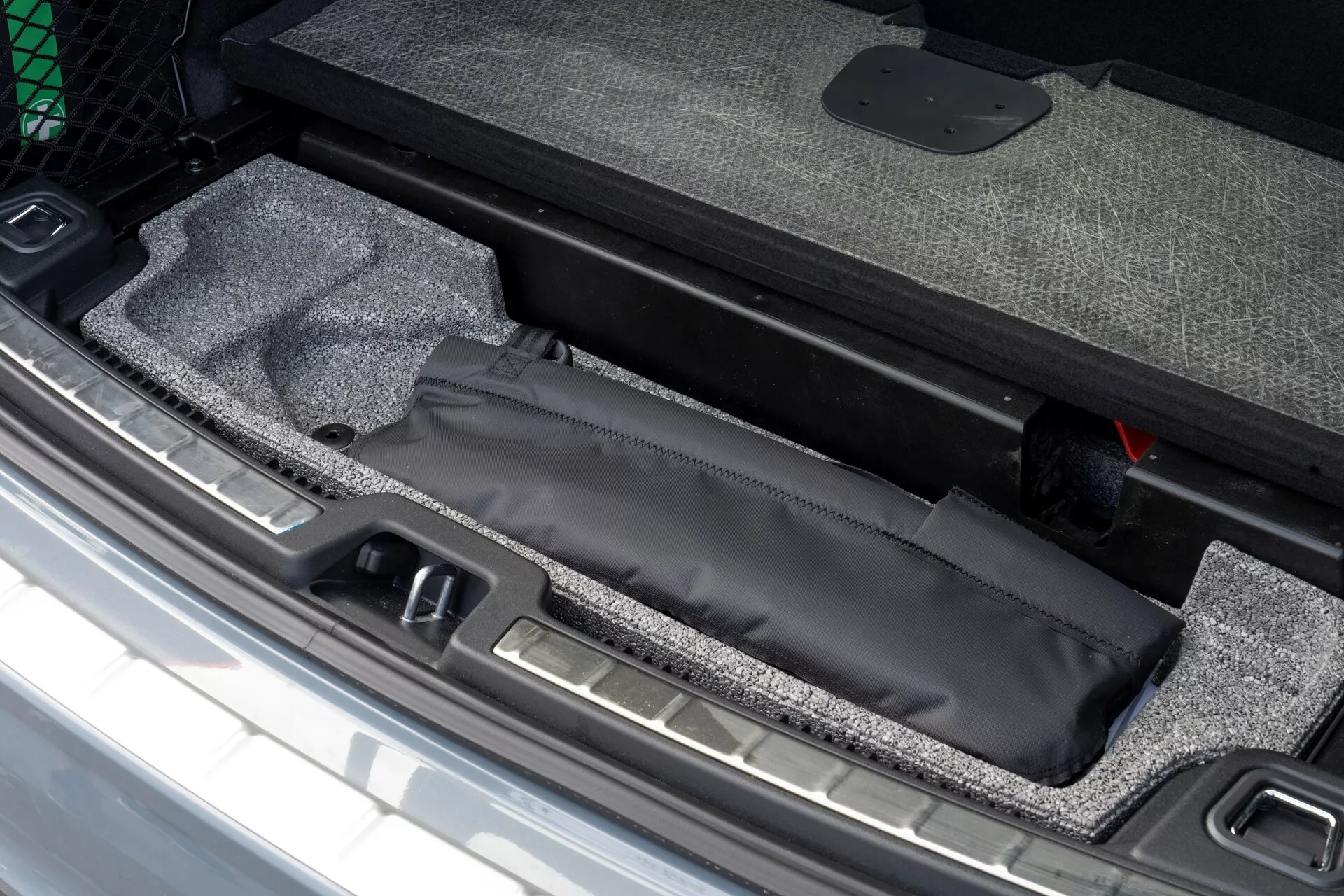 Primo piano dell'area di carico della Volvo XC60 2021 che mostra il vano portaoggetti nascosto sotto il pavimento del bagagliaio con custodia nera, interno rivestito in moquette e soglia in metallo visibile, accanto a uno strumento di emergenza rosso.