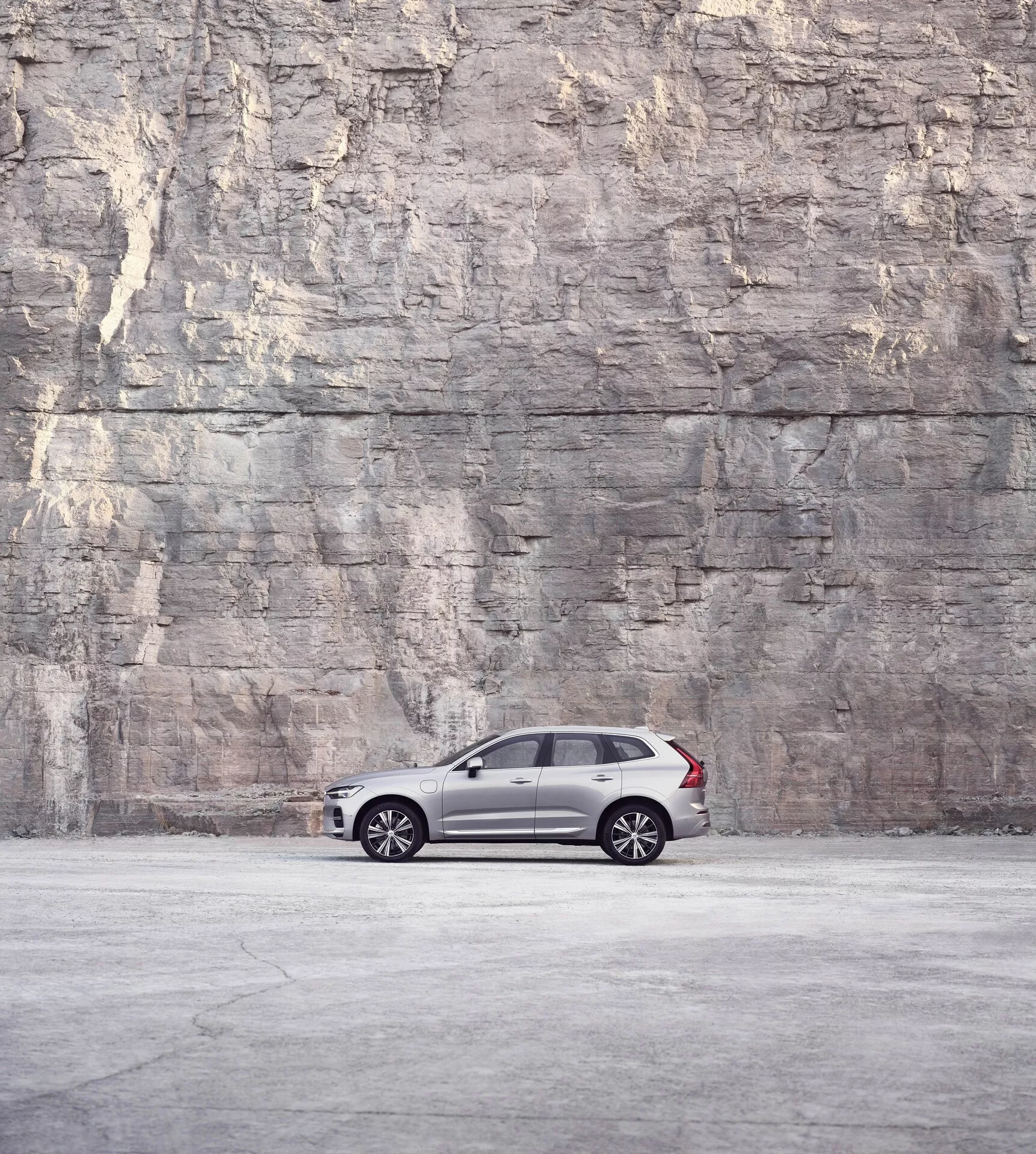 Vista laterale del profilo di una Volvo XC60 2021 (2ª generazione facelift) parcheggiata davanti a un grande muro di roccia o cemento texturizzato, che mette in mostra il suo design elegante e lussuoso con vernice metallica scura, cerchi a raggi multipli, finestrini oscurati e montanti B neri sotto una luce soffusa e diffusa.