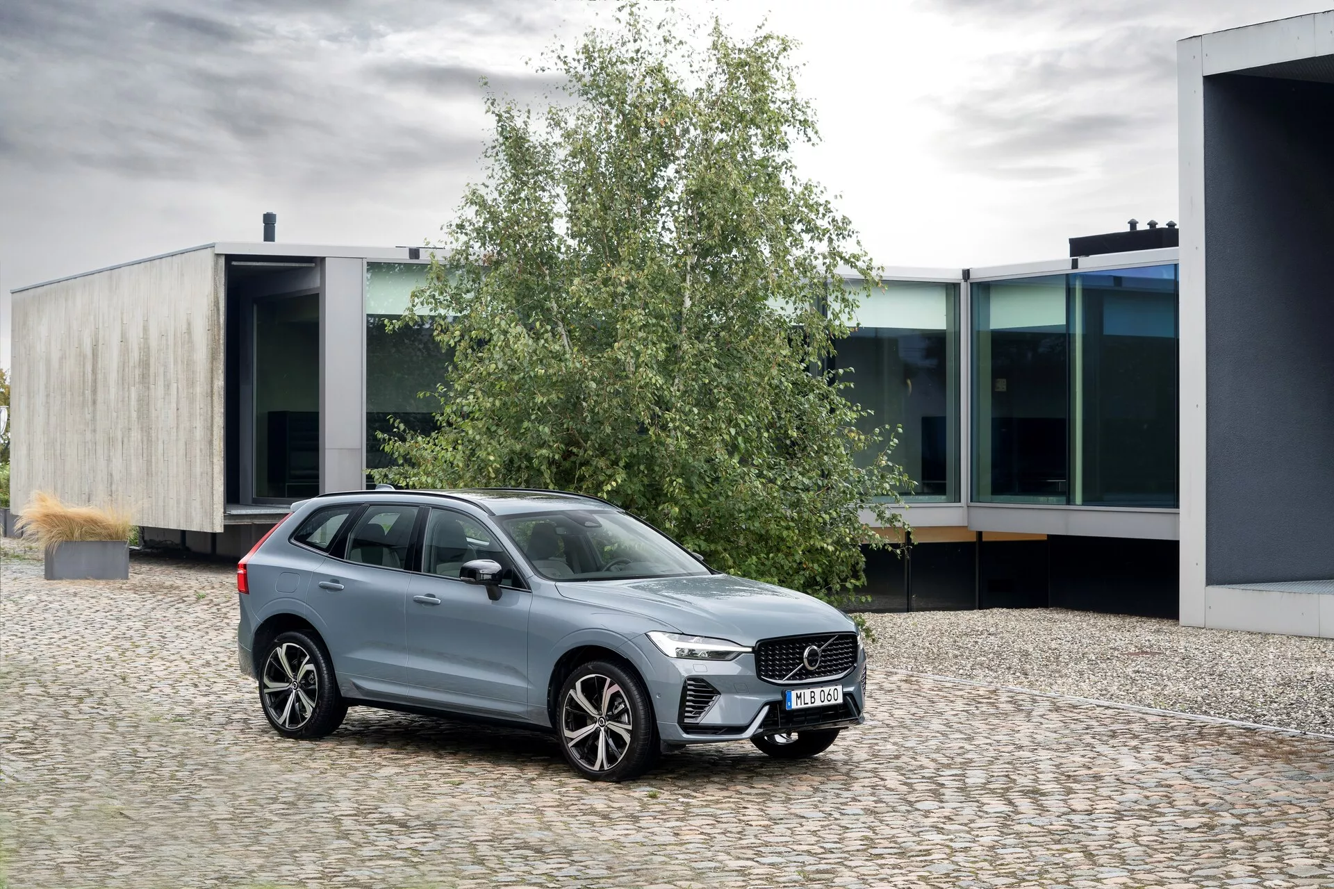 Immagine di una Volvo XC60 2021 (2ª generazione-restyling) parcheggiata su una superficie di ciottoli, posizionata ad un leggero angolo che guarda a sinistra. La prospettiva anteriore tre quarti mette in evidenza la griglia prominente con barre verticali, i fari LED eleganti con luci diurne e le grandi ruote in lega multiraggio. Le finestre sono bordate in cromo, migliorando la sua finitura metallica lucida. Il SUV presenta un'altezza da terra rialzata, barre sul tetto e specchietti laterali in tinta con la carrozzeria con indicatori di direzione integrati. La scena è ambientata in un contesto urbano con architettura moderna, evidenziando il suo design elegante e le caratteristiche di lusso.