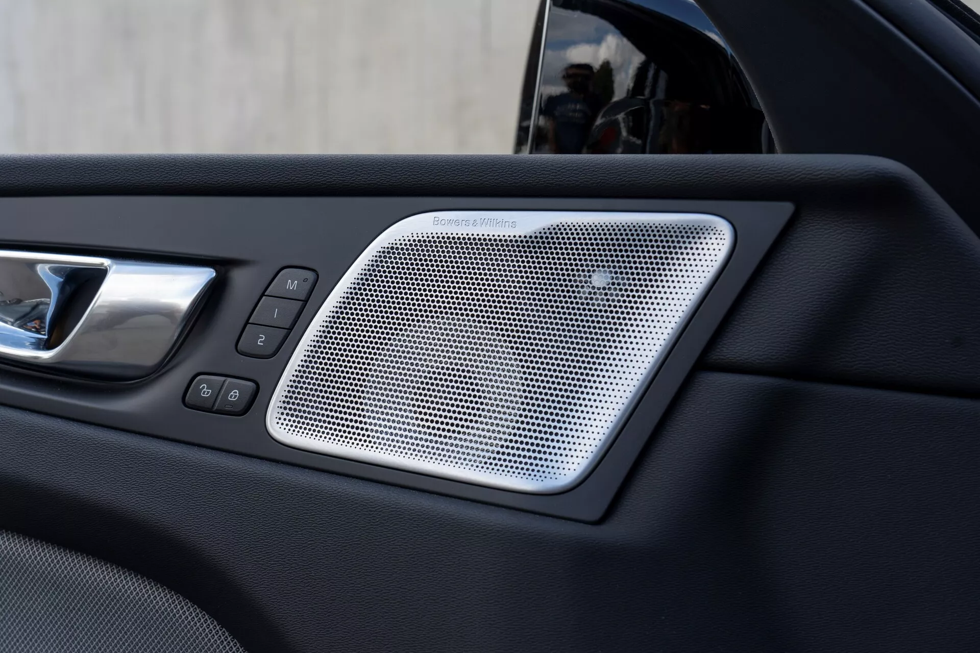 Immagine ravvicinata della sezione interna della portiera di una Volvo XC60 2021 (2ª generazione, restyling) che mostra una griglia dell'altoparlante Harman/Kardon con design a maglia, una maniglia della portiera cromata e comandi per finestrini e serrature elettriche, enfatizzando il lusso e la modernità delle caratteristiche del veicolo.