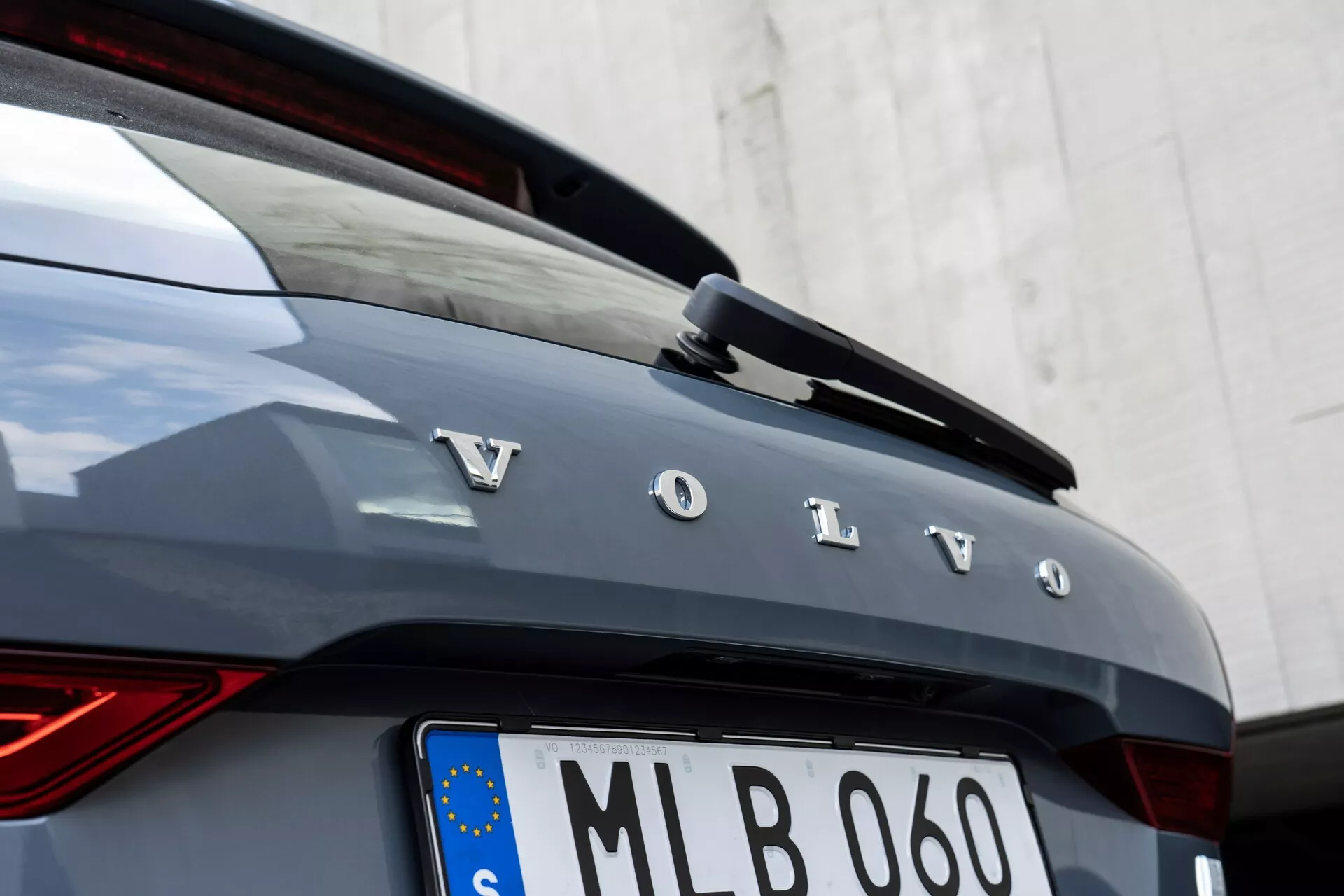 Vista dettagliata della parte posteriore di un'auto Volvo XC60 2021 (seconda generazione facelift). L'immagine mostra lettere argentate e in grassetto che formano la scritta 'Volvo' sul portellone posteriore, parte del lunotto posteriore e uno spoiler posteriore integrato con una terza luce di stop a LED. L'auto è di colore grigio scuro o grafite, con una luce posteriore LED verticale sul lato sinistro che presenta un design a 'martello di Thor'. Sotto il nome del marchio è presente una targa europea con una bandiera dell'UE, che indica la registrazione nell'UE ed è incorniciata da un bordo cromatico. L'angolo della telecamera è leggermente spostato a destra, focalizzandosi sulle lettere e sui bordi della carrozzeria dell'auto, con uno sfondo bianco neutro.