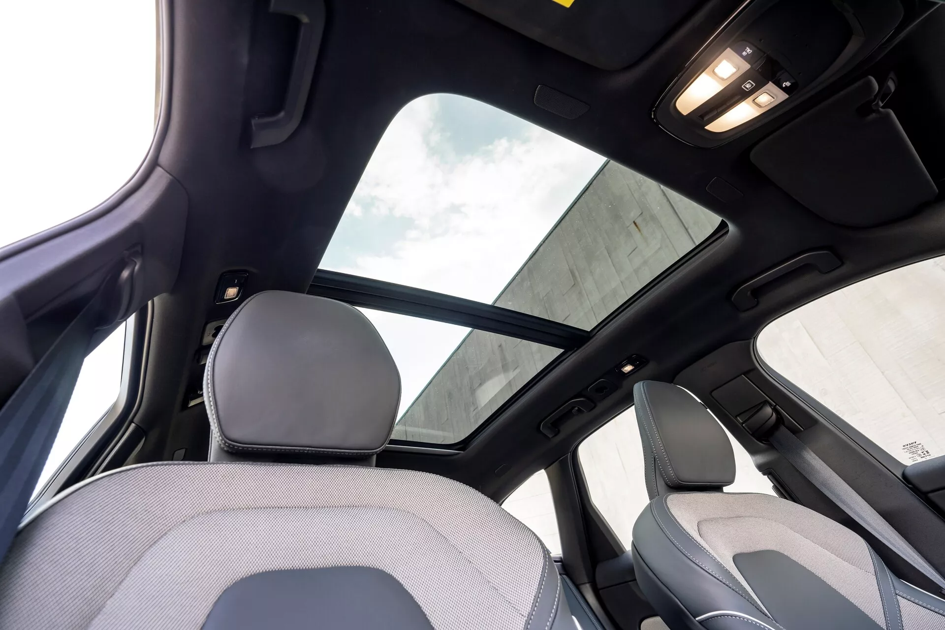 Interni della Volvo XC60 2021 restyling seconda generazione visti dal sedile posteriore, concentrandosi sulla sezione superiore dell'abitacolo. Caratteristiche notevoli includono un grande tetto panoramico con parasole, rivestimento del tetto nero che indica un alto livello di allestimento o un pacchetto di lusso, e illuminazione interna sul soffitto. L'immagine cattura parte del poggiatesta e dello schienale superiore del sedile del passeggero anteriore, inclusi il montante B e la maniglia sopra la porta posteriore.