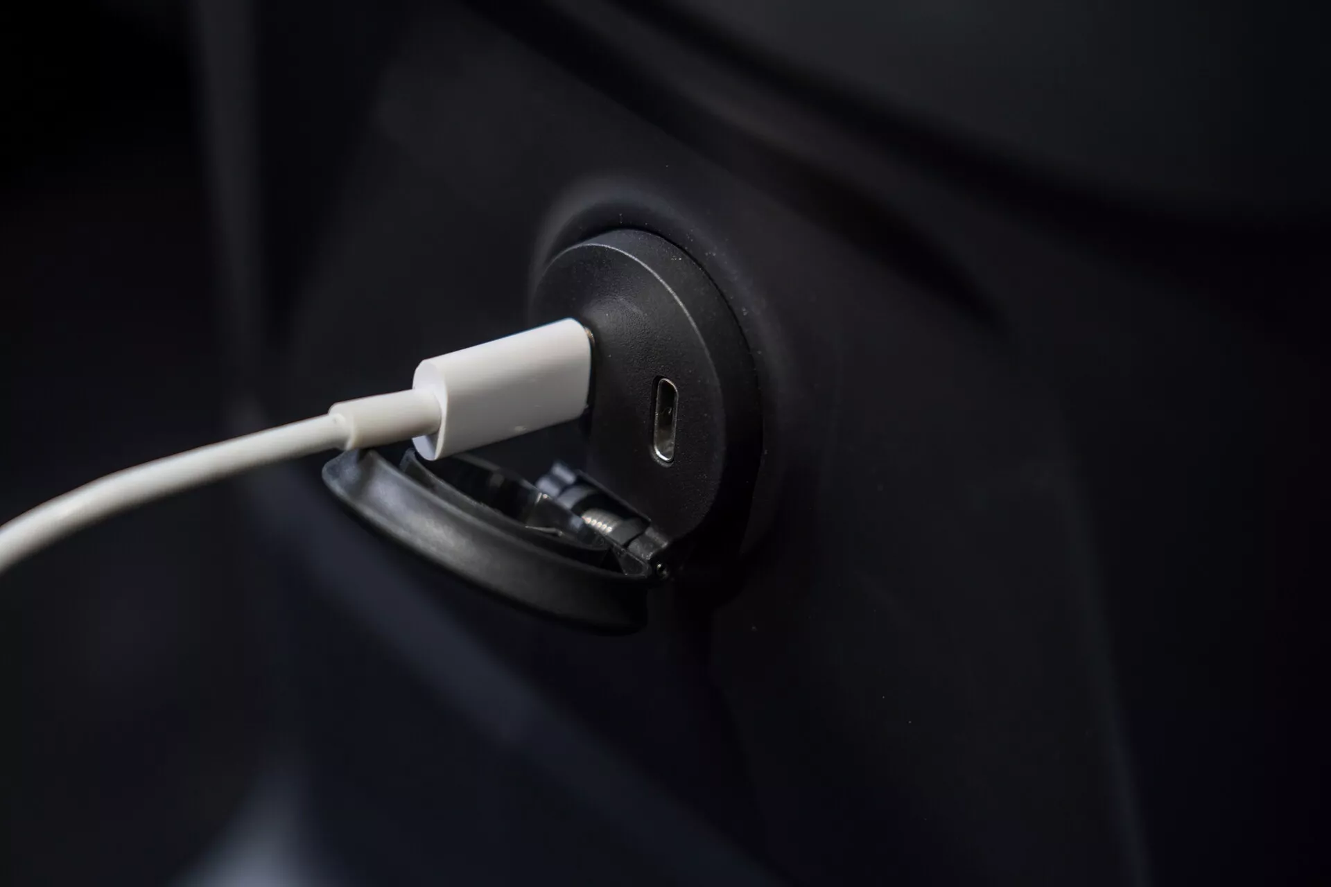 Primo piano del porto di ricarica del veicolo elettrico Volvo XC60 2021 con il cavo collegato, che sottolinea il design elegante e moderno. L'inquadratura è stretta sul porto di ricarica, lasciando fuori vista la maggior parte del corpo scuro del veicolo. Lo sfondo sfocato mette in risalto la precisione del processo di ricarica EV, simboleggiando la tecnologia verde e il trasporto sostenibile.